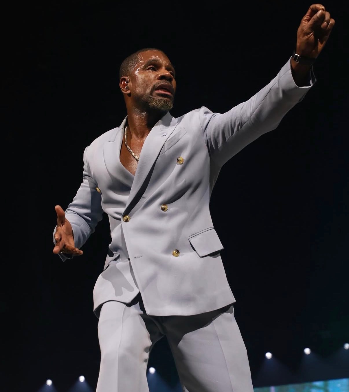 Man in a light gray suit gesturing with his right arm raised, on a dark stage with lights, serious expression.