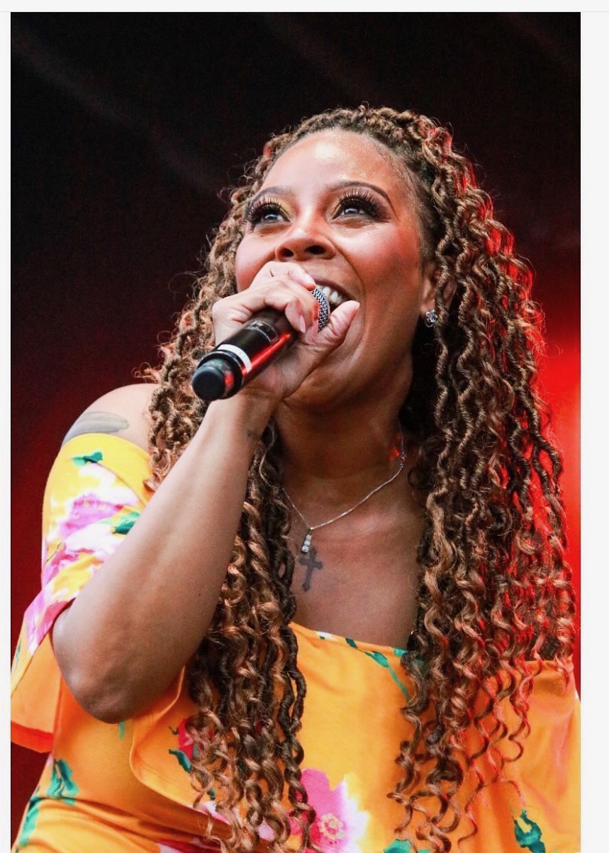 Woman singing into microphone with curly hair, wearing a yellow dress with colorful patterns, smiling brightly.