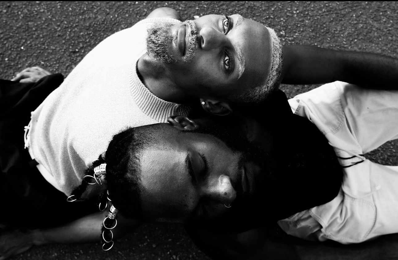 Two people lying on the ground with heads close together, one looking up and the other with eyes closed.
