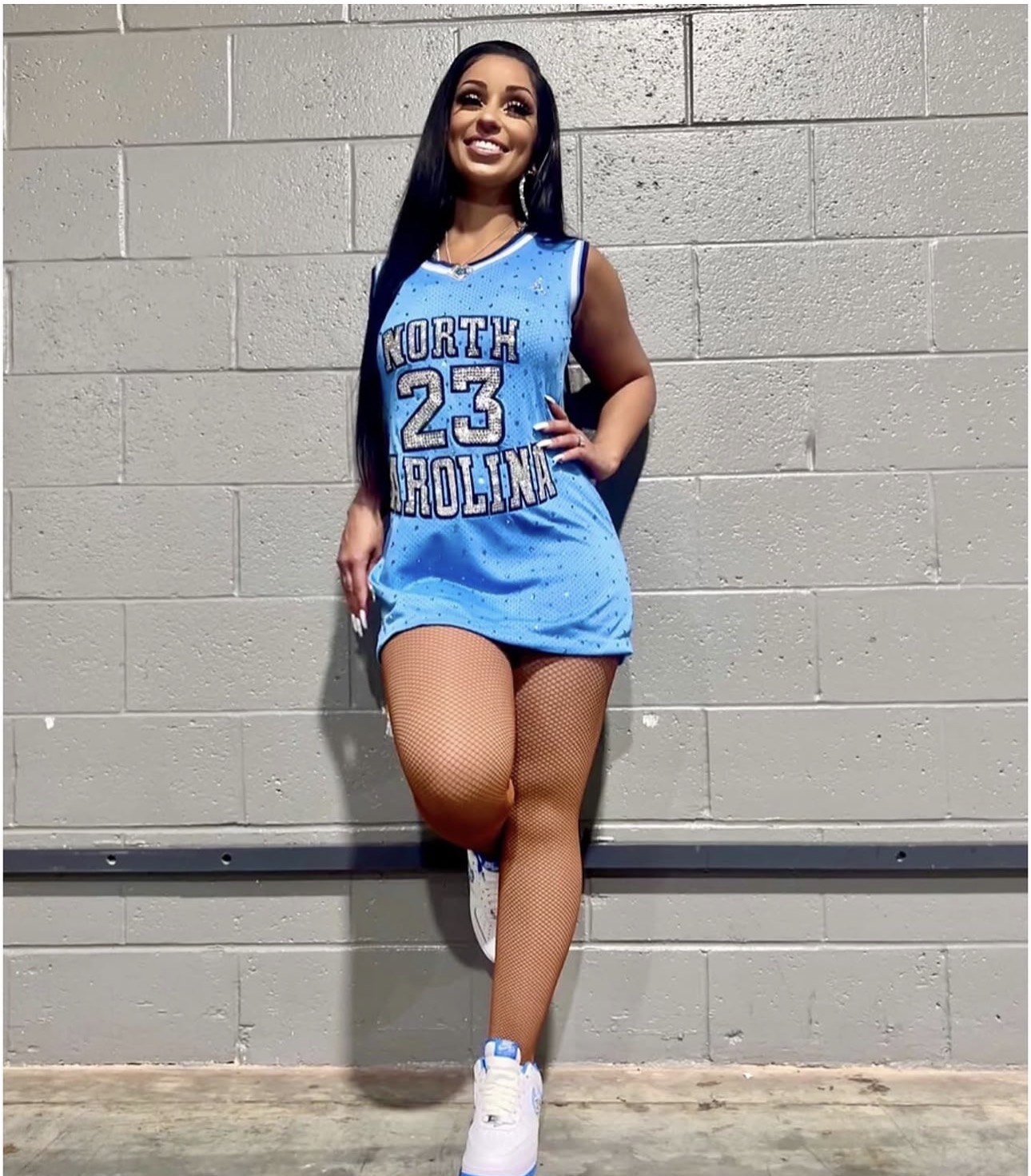 Young woman in a blue basketball jersey with the number 23, standing against a gray brick wall, smiling, with one hand on her hip.