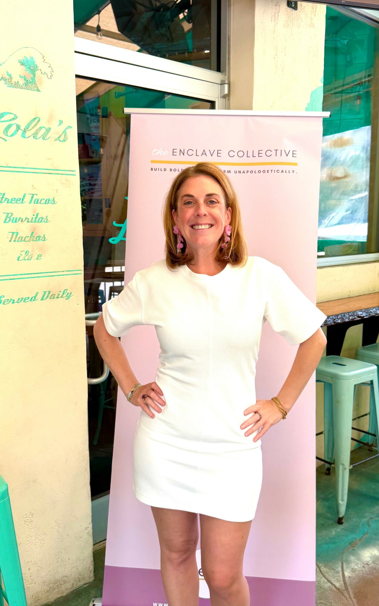 Woman in white dress standing indoors with hands on hips, smiling, behind a pink banner, in a casual setting.