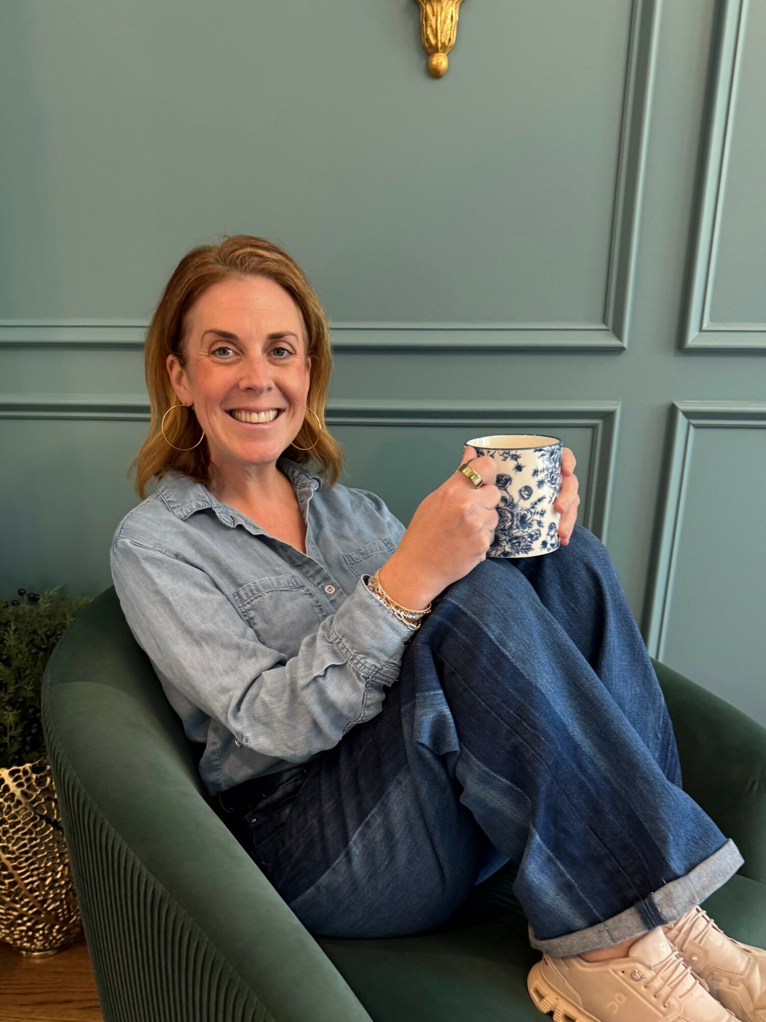 Woman sitting in a green chair holding a mug, smiling, against a teal wall with decorative paneling.