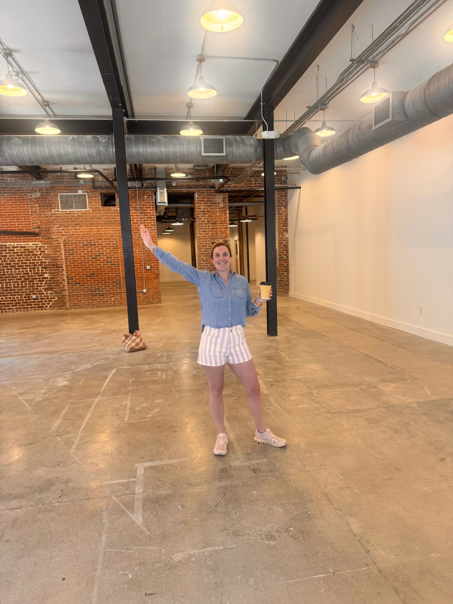 Person standing in an indoor space with arms outstretched, wearing a blue jacket and striped skirt, with brick walls and ceiling lights.
