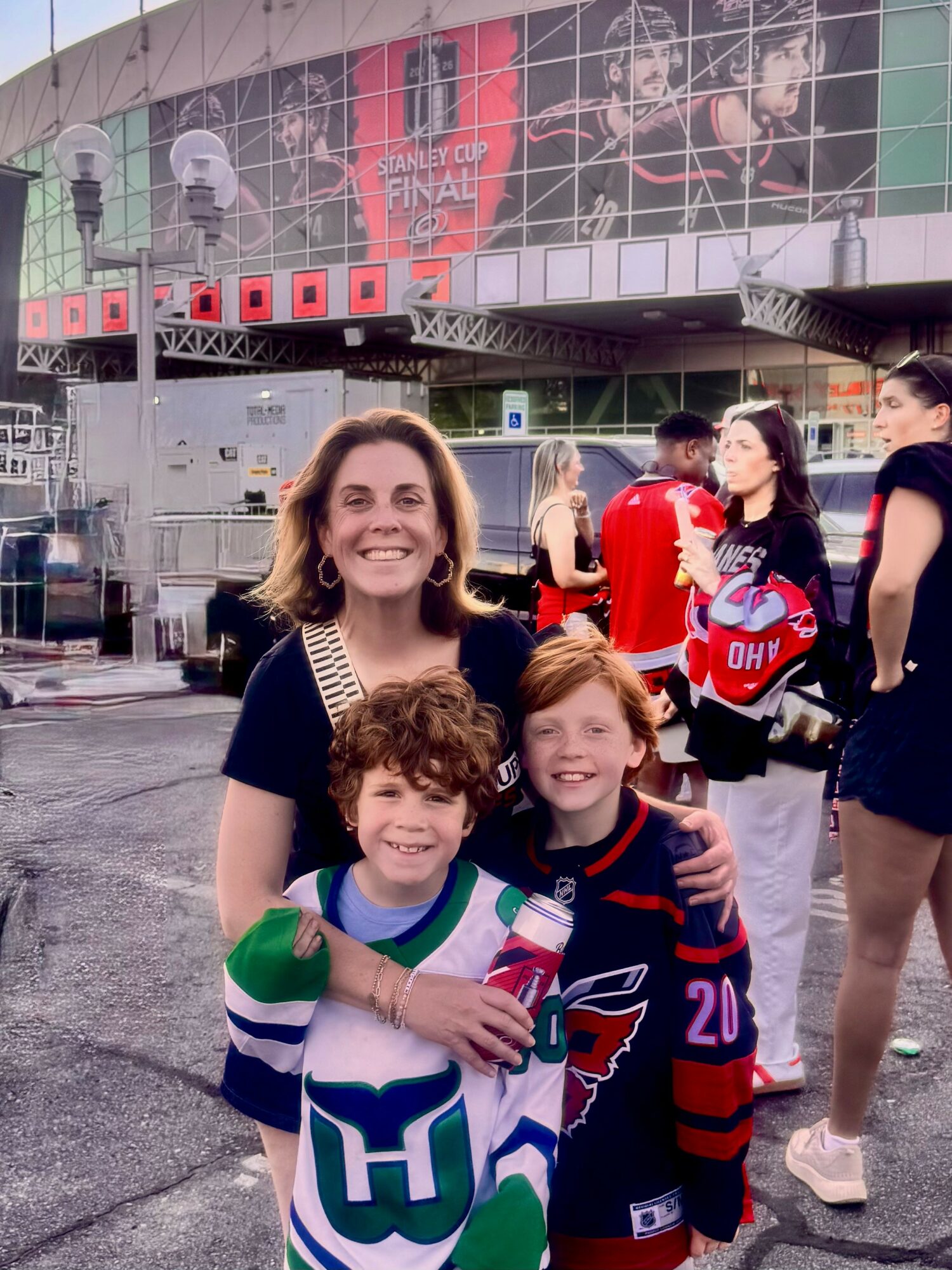 Smiling woman with two children in hockey jerseys outdoors, with a stadium and people in the background.