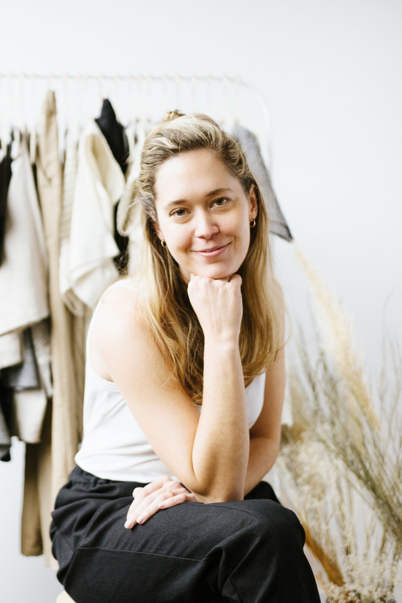 Young woman with long blonde hair sitting with her chin resting on her hand, smiling, in front of clothing racks.