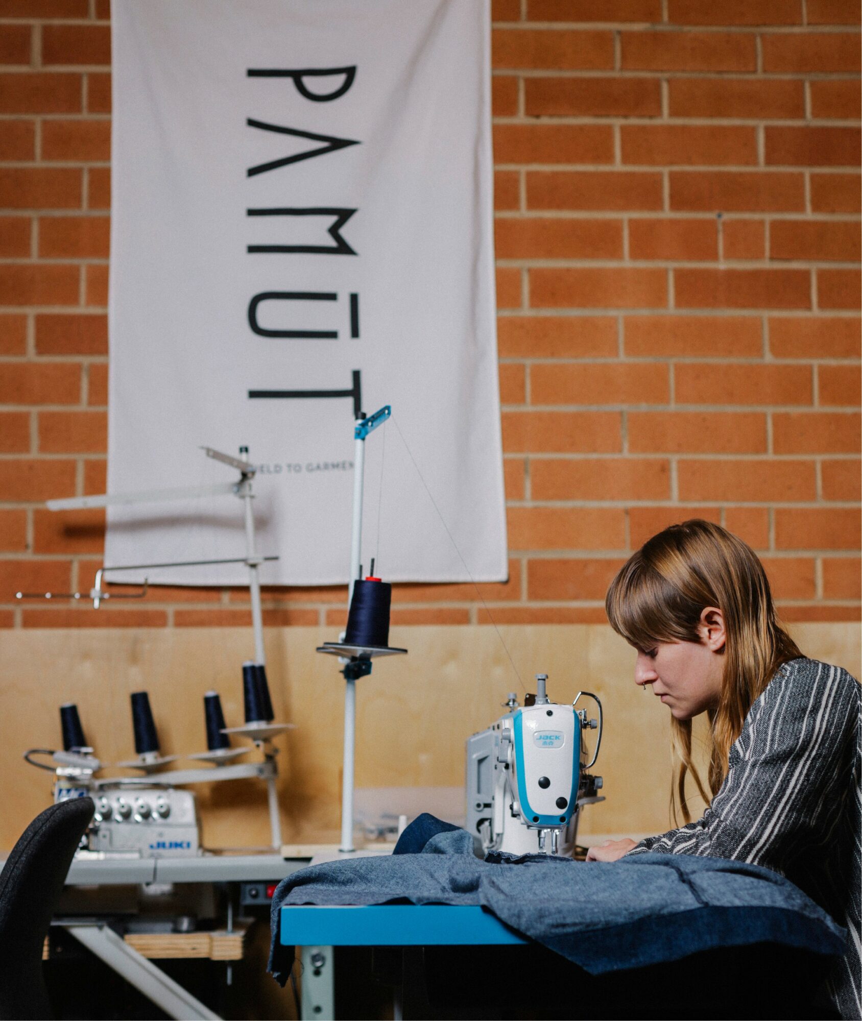 Person sewing fabric on a machine in a workshop with a brick wall and a banner reading 'PYMUT'.