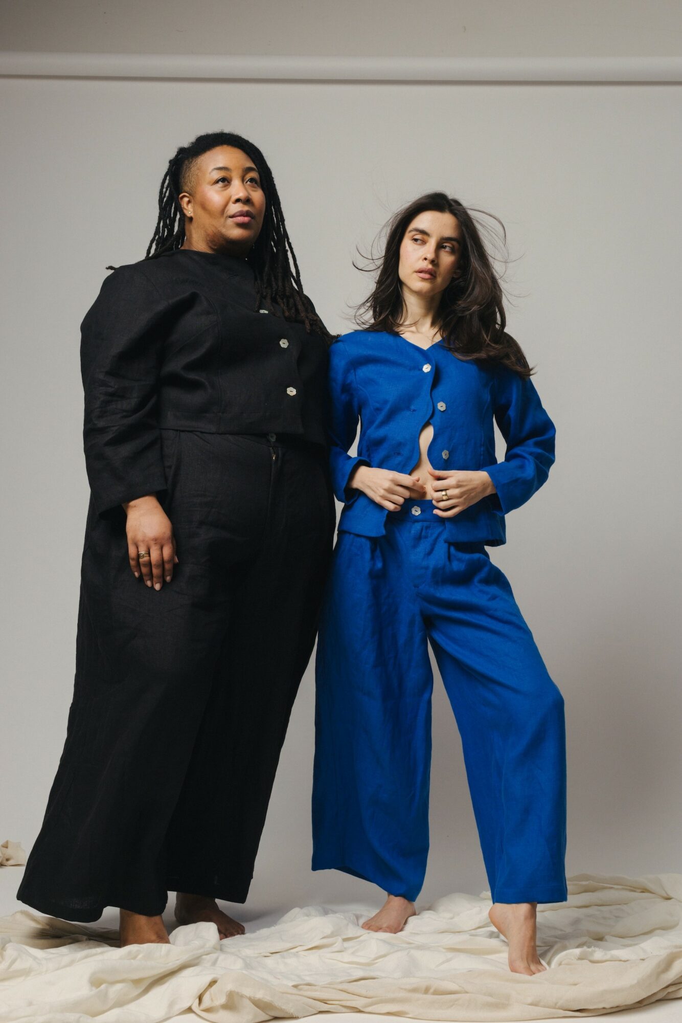 Two women stand side by side against a plain background, one in black and the other in blue, both looking at the camera.