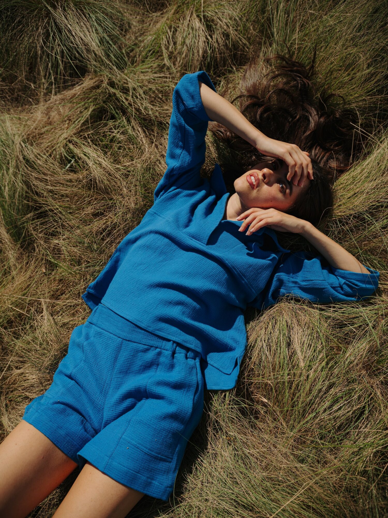 Child lying on grass with hand over eyes, wearing a blue outfit, eyes closed, outdoors.