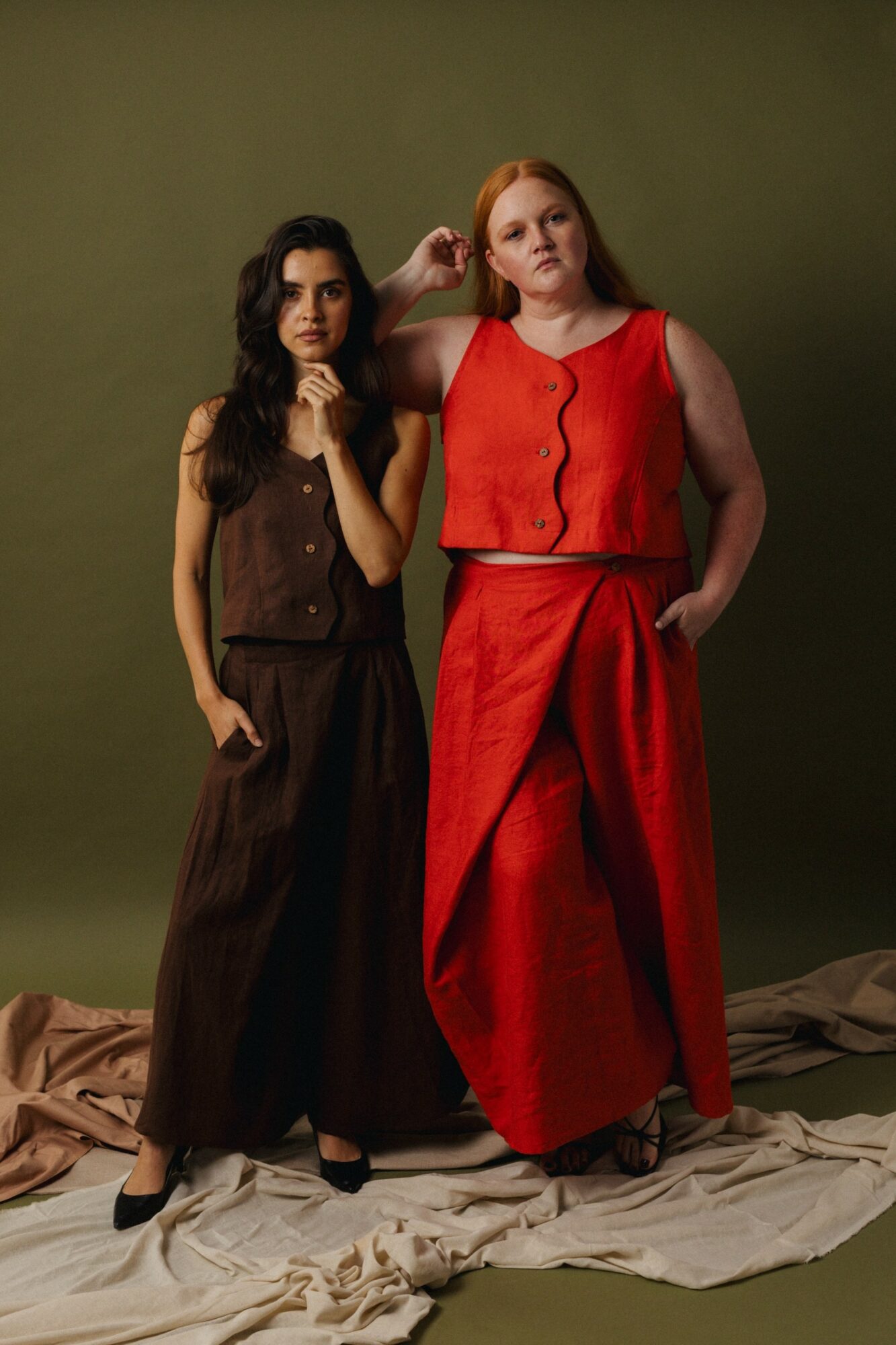 Two women stand side by side against a green background, one in a brown outfit and the other in a red outfit, with fabric on the floor.