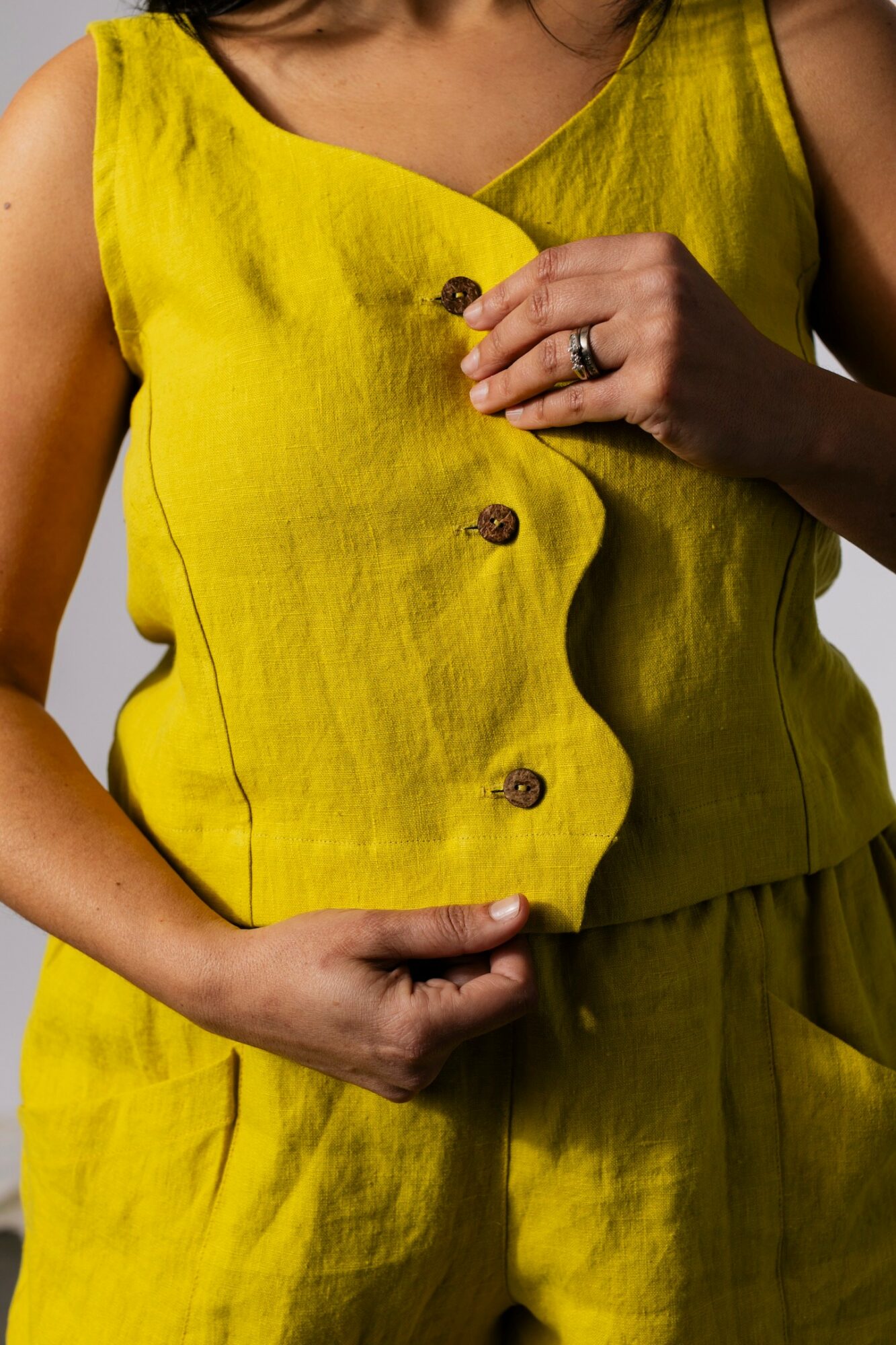 Person wearing a yellow sleeveless top with buttons, holding the fabric near the waist, with a ring on finger.