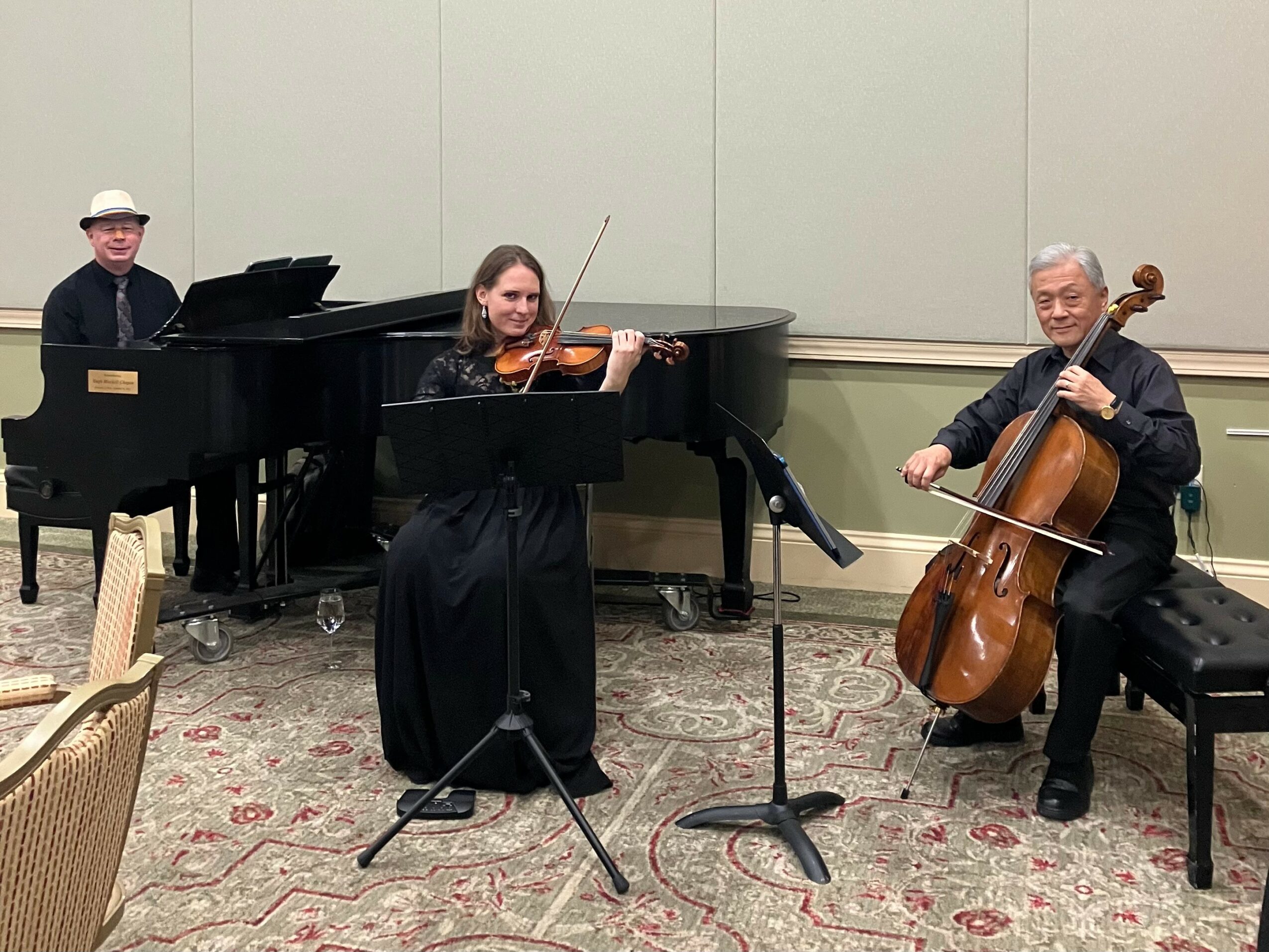 Three musicians perform with a piano, violin, and cello in a room with a patterned carpet and plain wall.