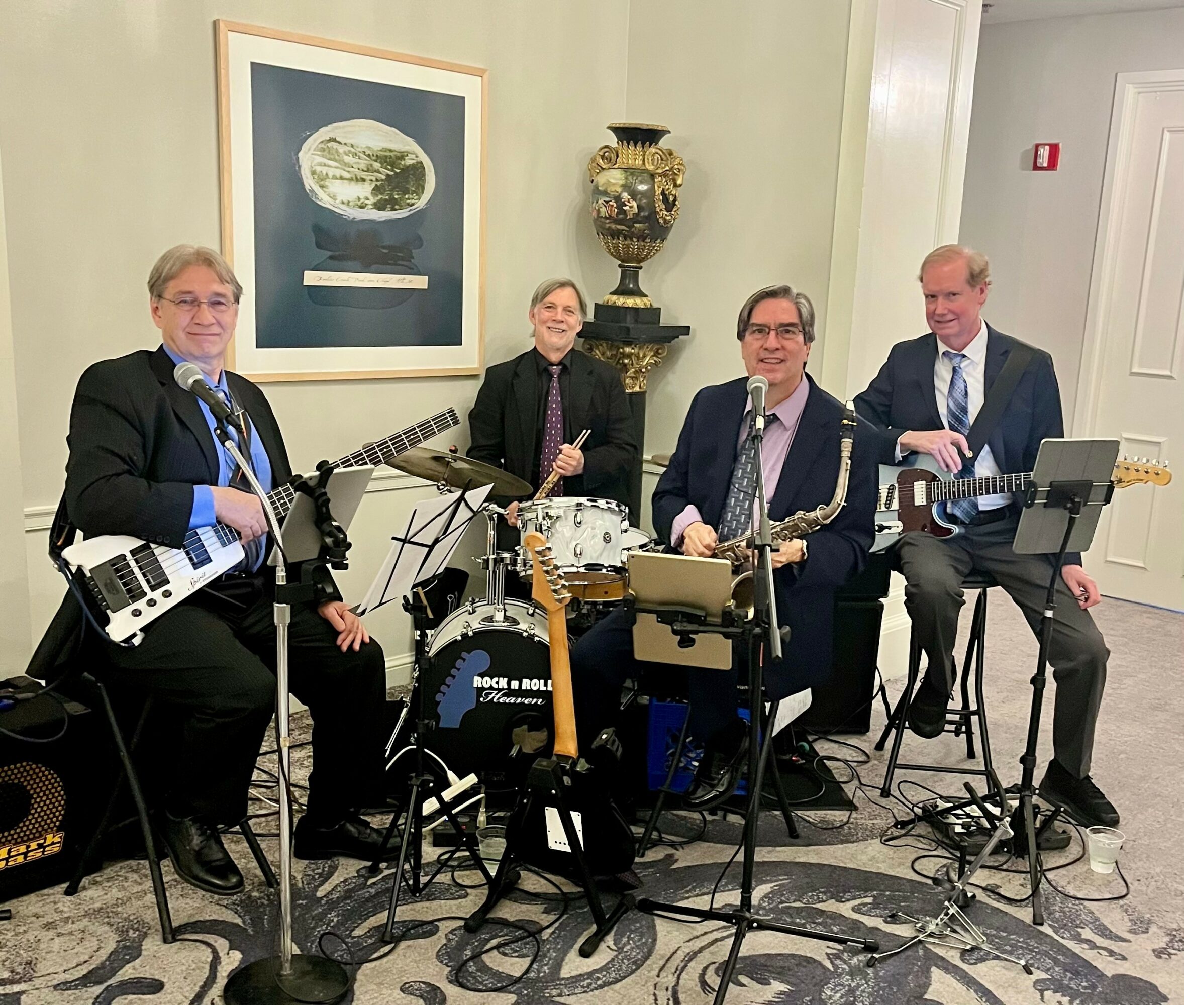 Four men in suits with musical instruments, standing in a room with artwork and a decorative vase, preparing to perform.