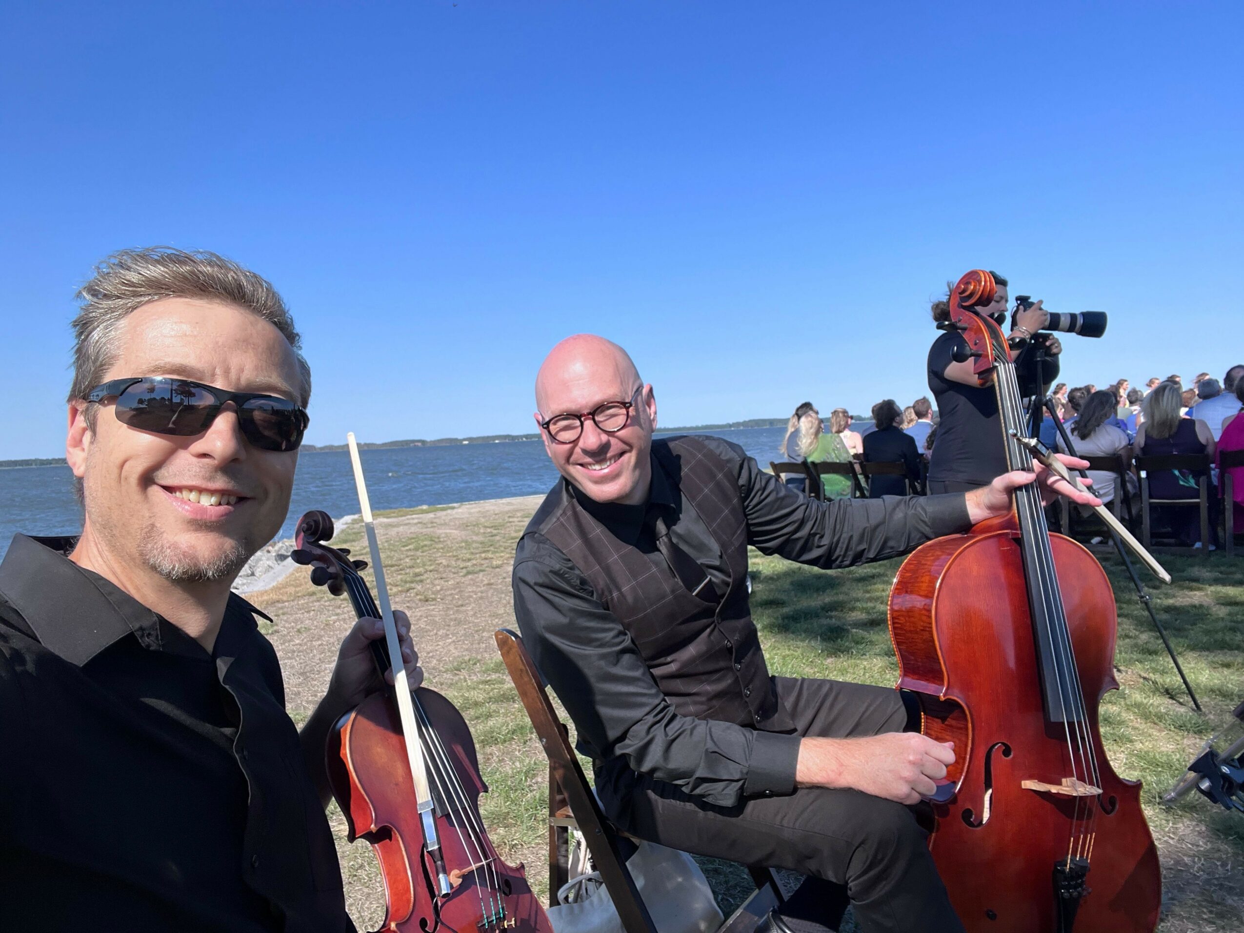 Two musicians with violins outdoors, one smiling and wearing sunglasses, the other playing violin, with a clear blue sky.