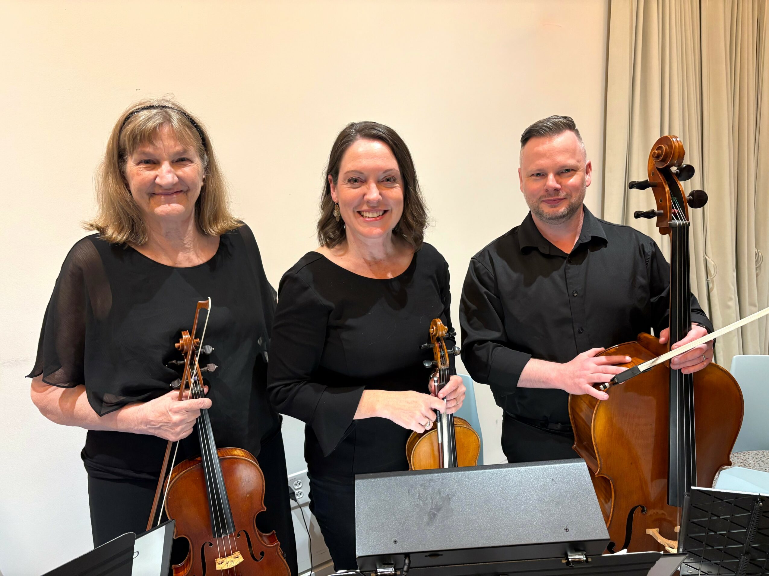 Three musicians holding violins and a viola, standing indoors with a cream-colored wall and curtains behind them.