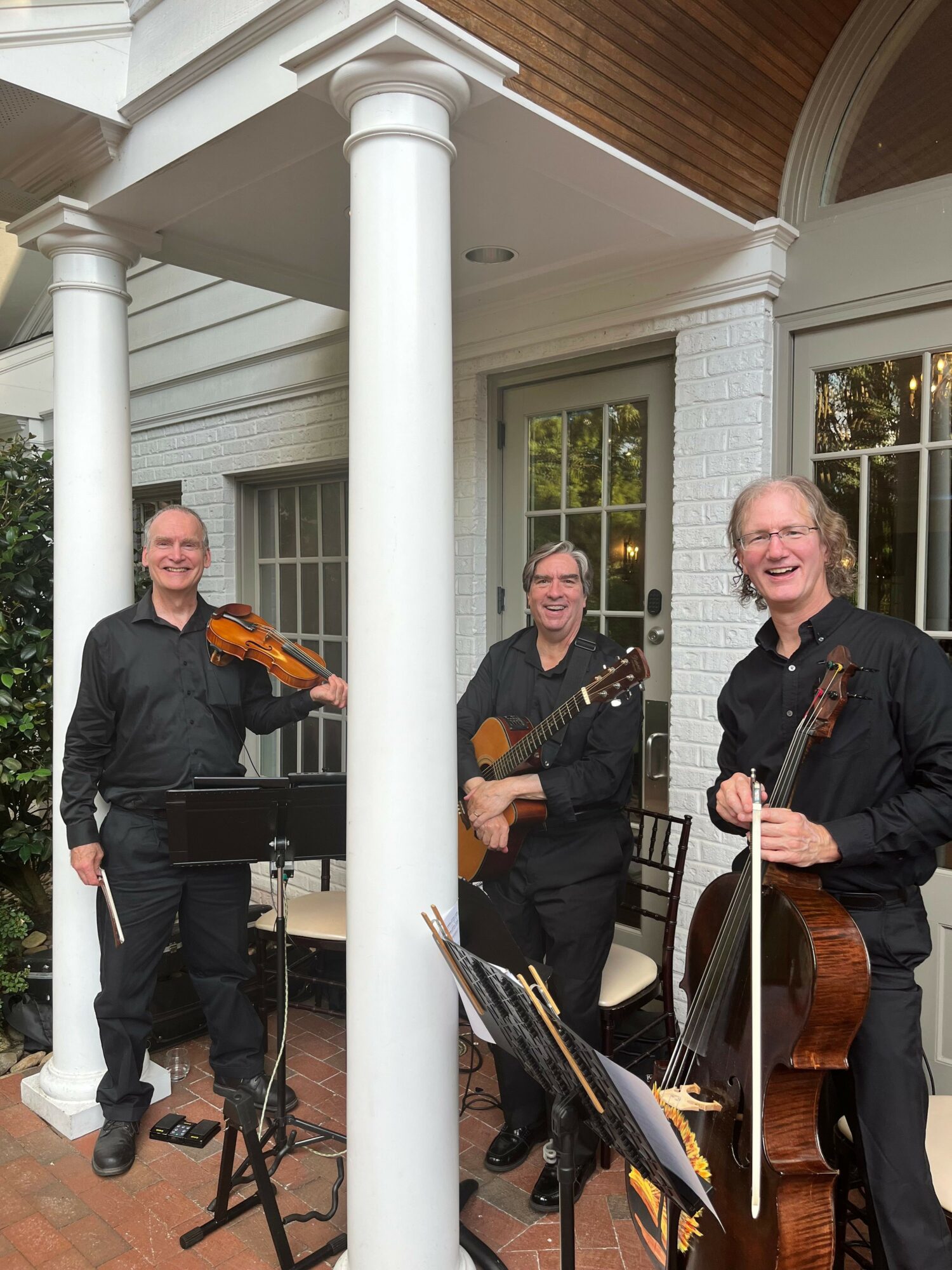 Two musicians with violins and a cello in a room with large windows and white columns.