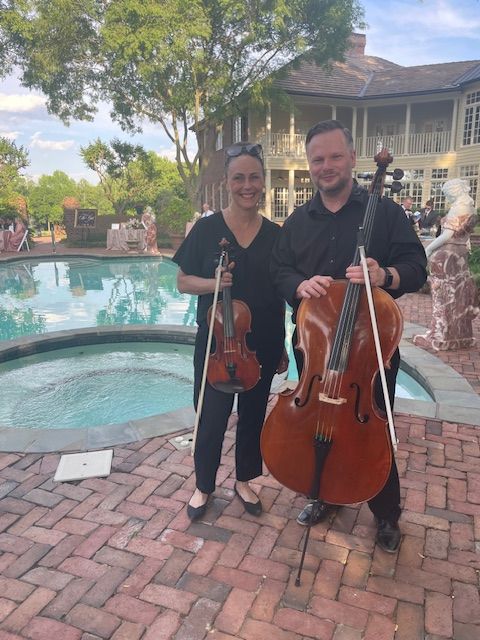 Two musicians, one holding a cello and the other a violin, standing outdoors near a fountain with trees and buildings in the background.