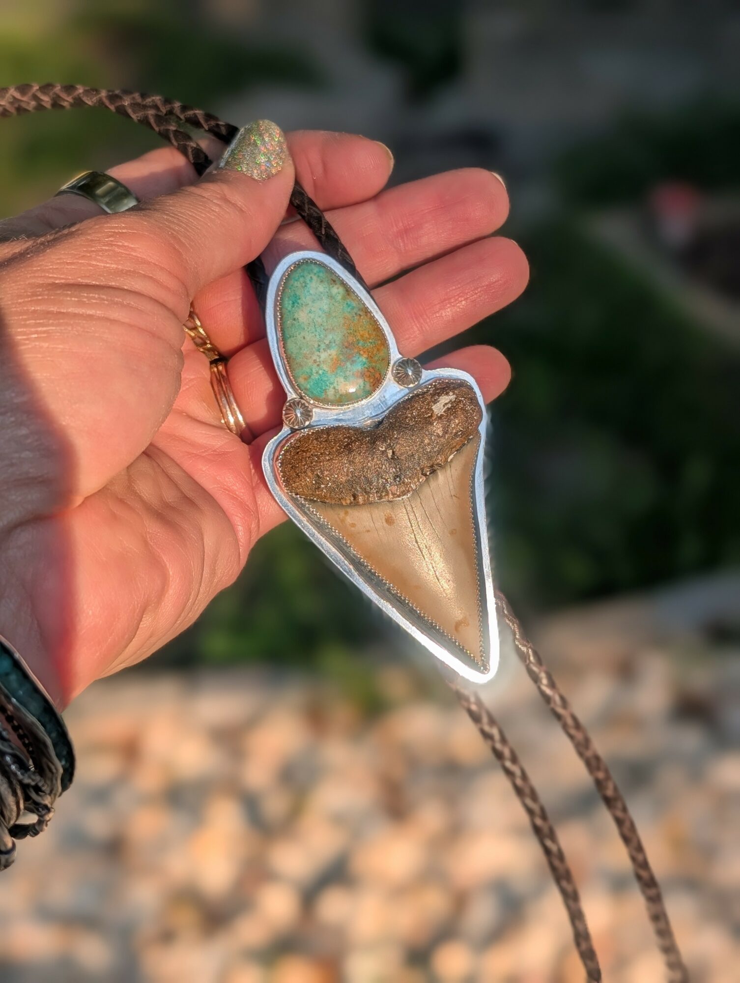 Hand holding a pendant with a green and blue stone and a brown, glittery heart-shaped element, outdoors with blurred background.