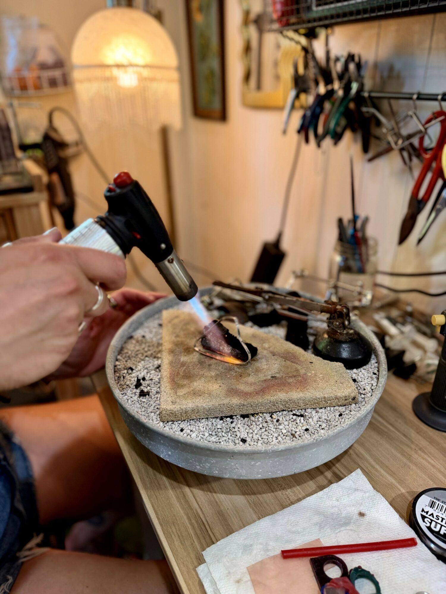 Person using a torch to work on a small object on a stone platform in a workshop, with tools hanging on the wall.
