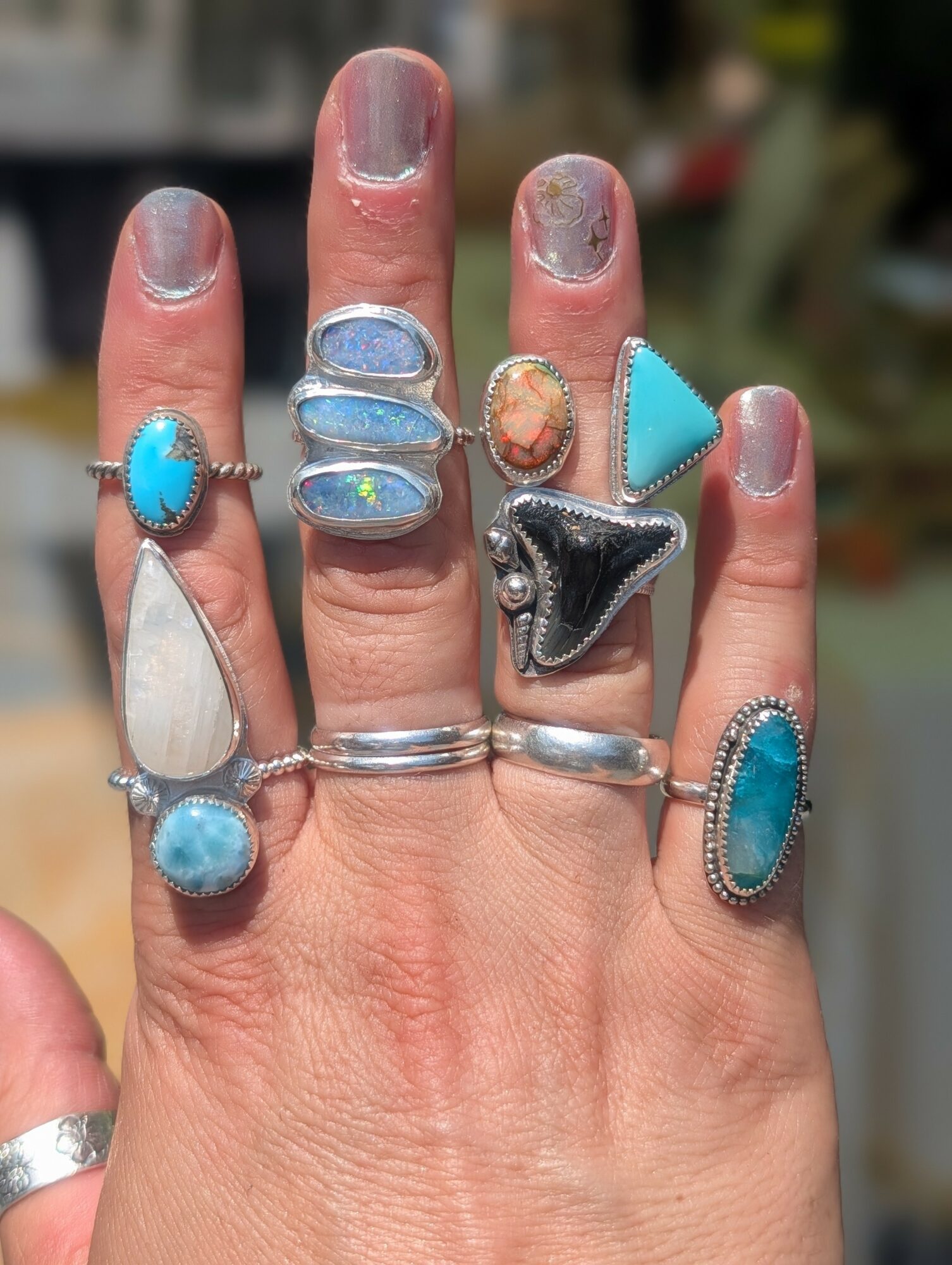 Hand with multiple rings featuring turquoise, silver, and colorful stones, against a blurred outdoor background.