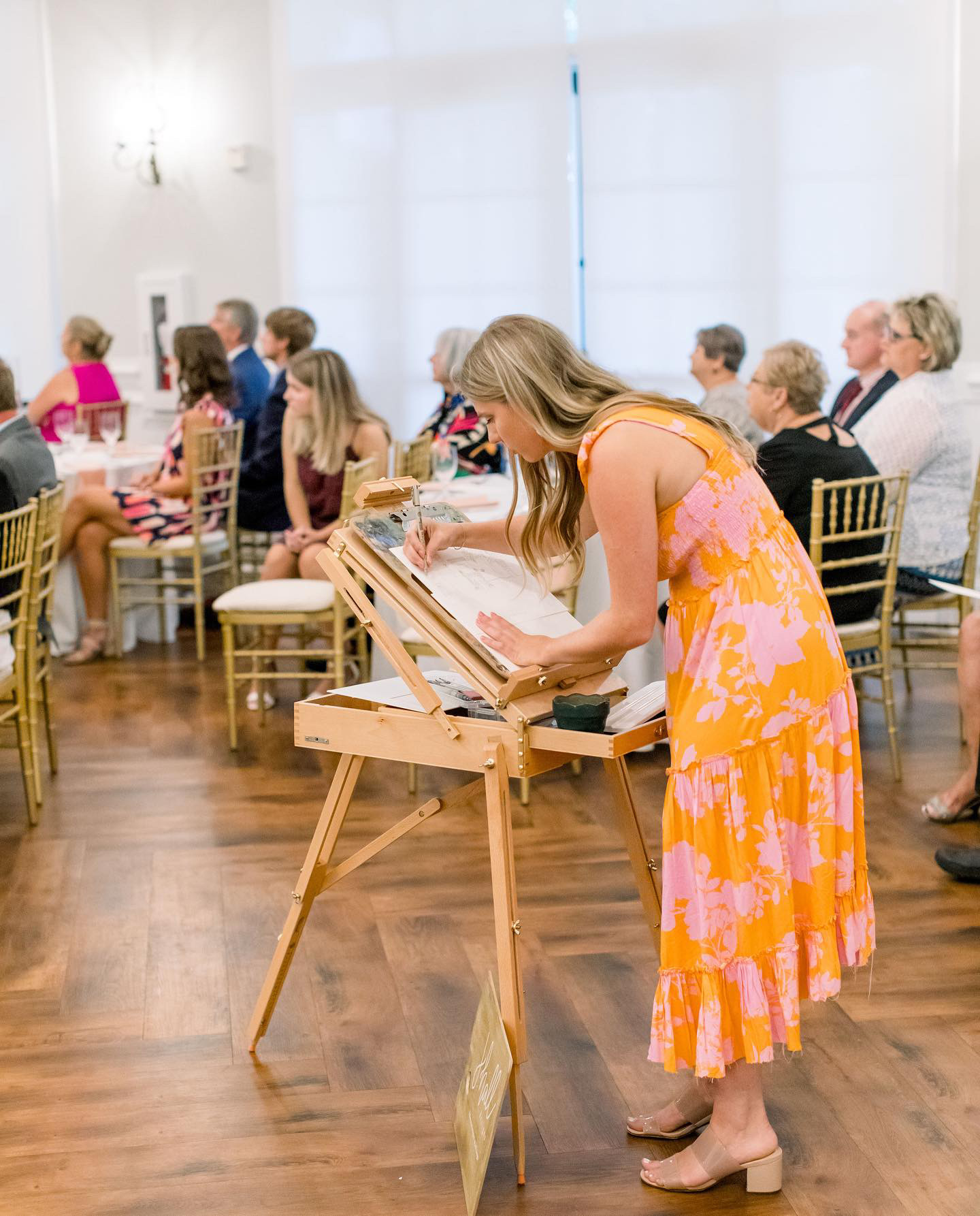 Woman in a pink and orange dress reads a book at a wooden stand in a room with seated audience members, wooden floor, and white curtains.