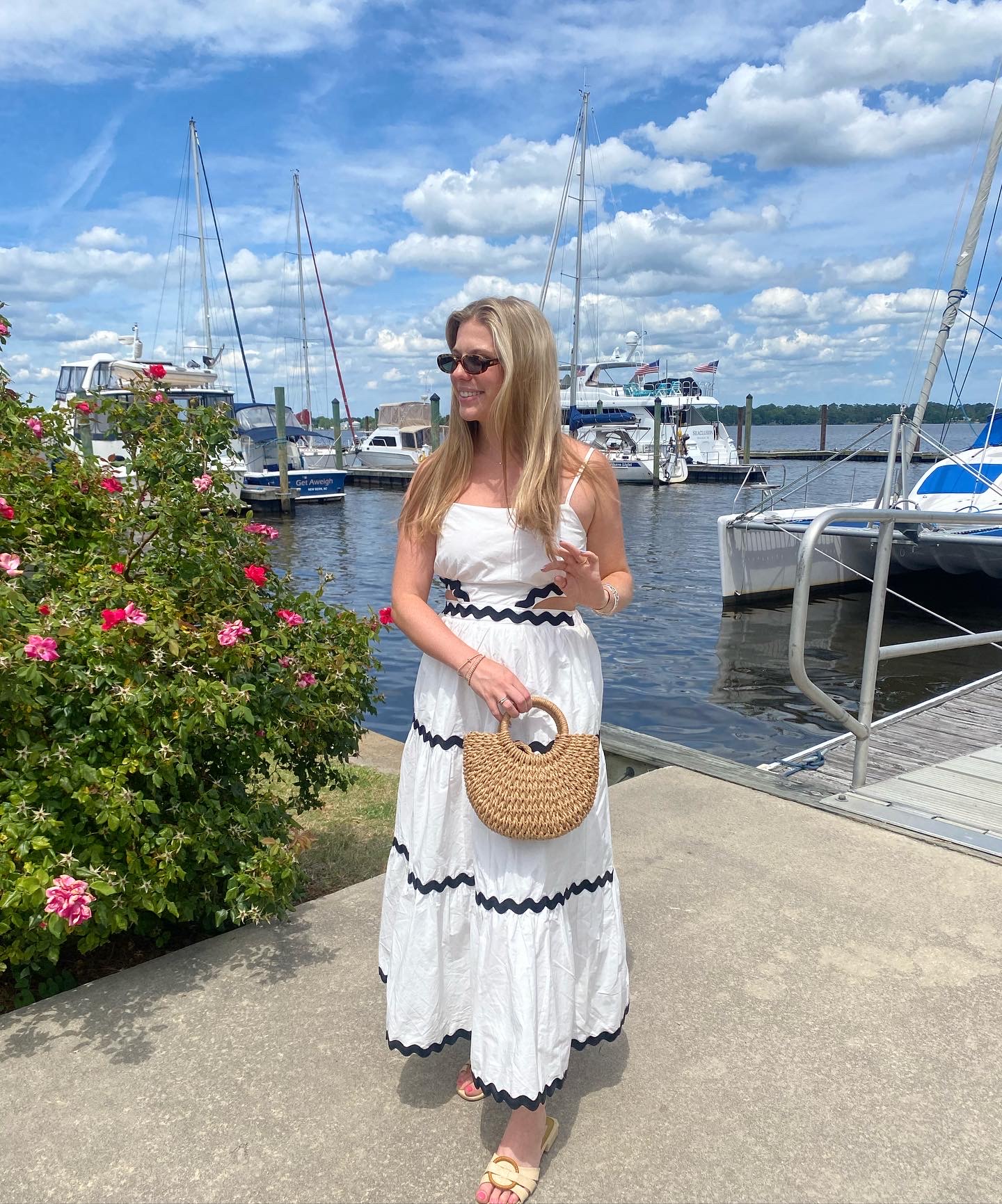Woman in a white dress with black stripes standing near a marina with boats and a blue sky.