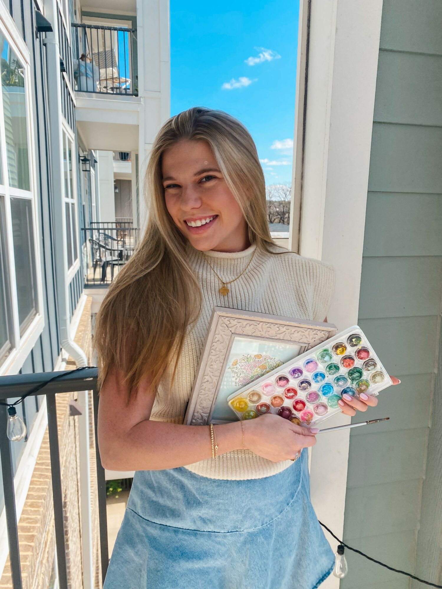 Young woman with long hair smiling, holding a watercolor set and a sketchbook, standing near a window with a blue sky background.
