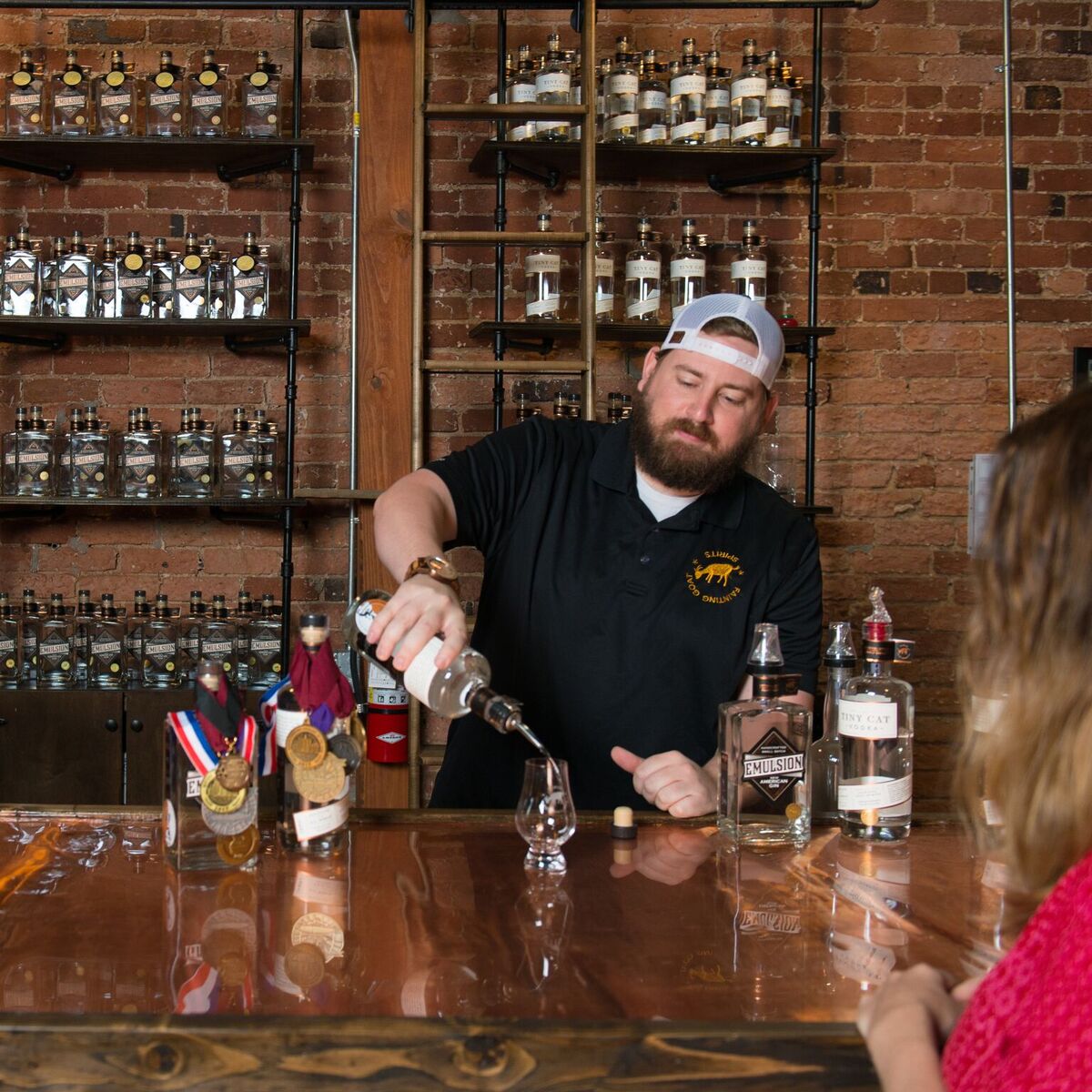 Bartender with a beard and white cap pours liquid into a glass at a bar counter, shelves with bottles behind him.