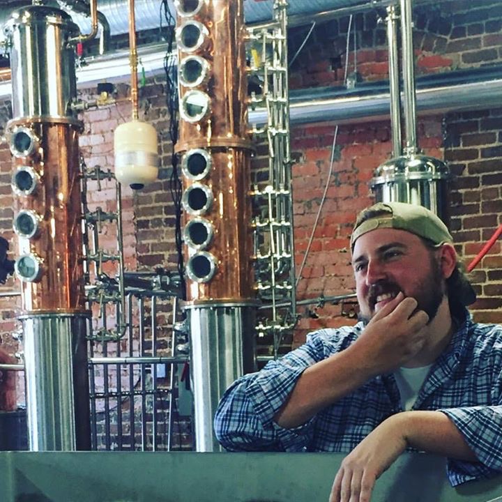 Man with a beard and cap smiling, resting his chin on his hand, in front of copper distillation equipment, brick wall background.