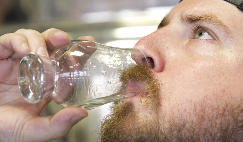 Man with beard drinking from a glass bottle, close-up of face and hand, neutral background.