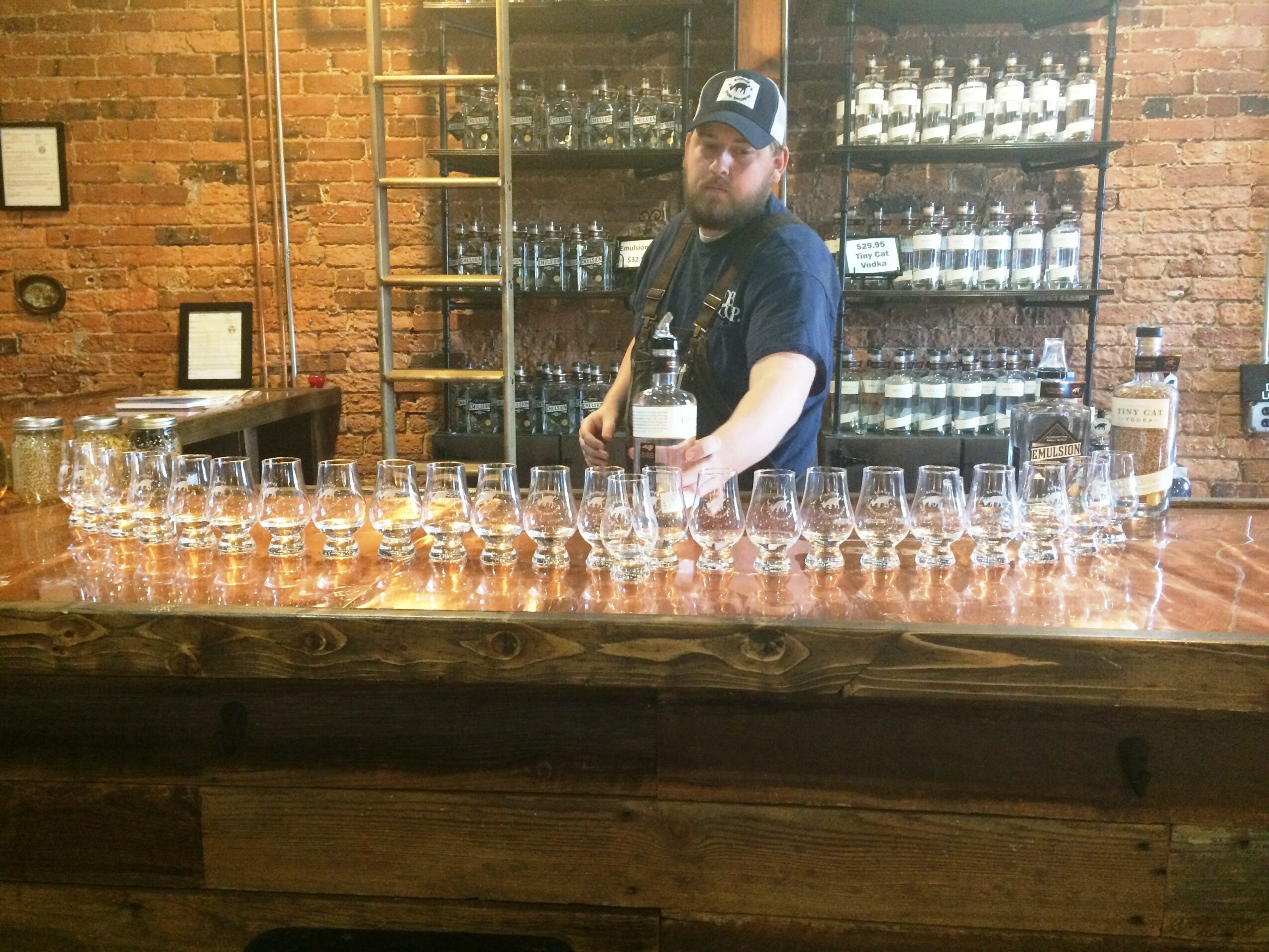 Man with beard and cap pours drink behind bar with glasses, brick wall background, shelves with bottles, wooden bar counter.