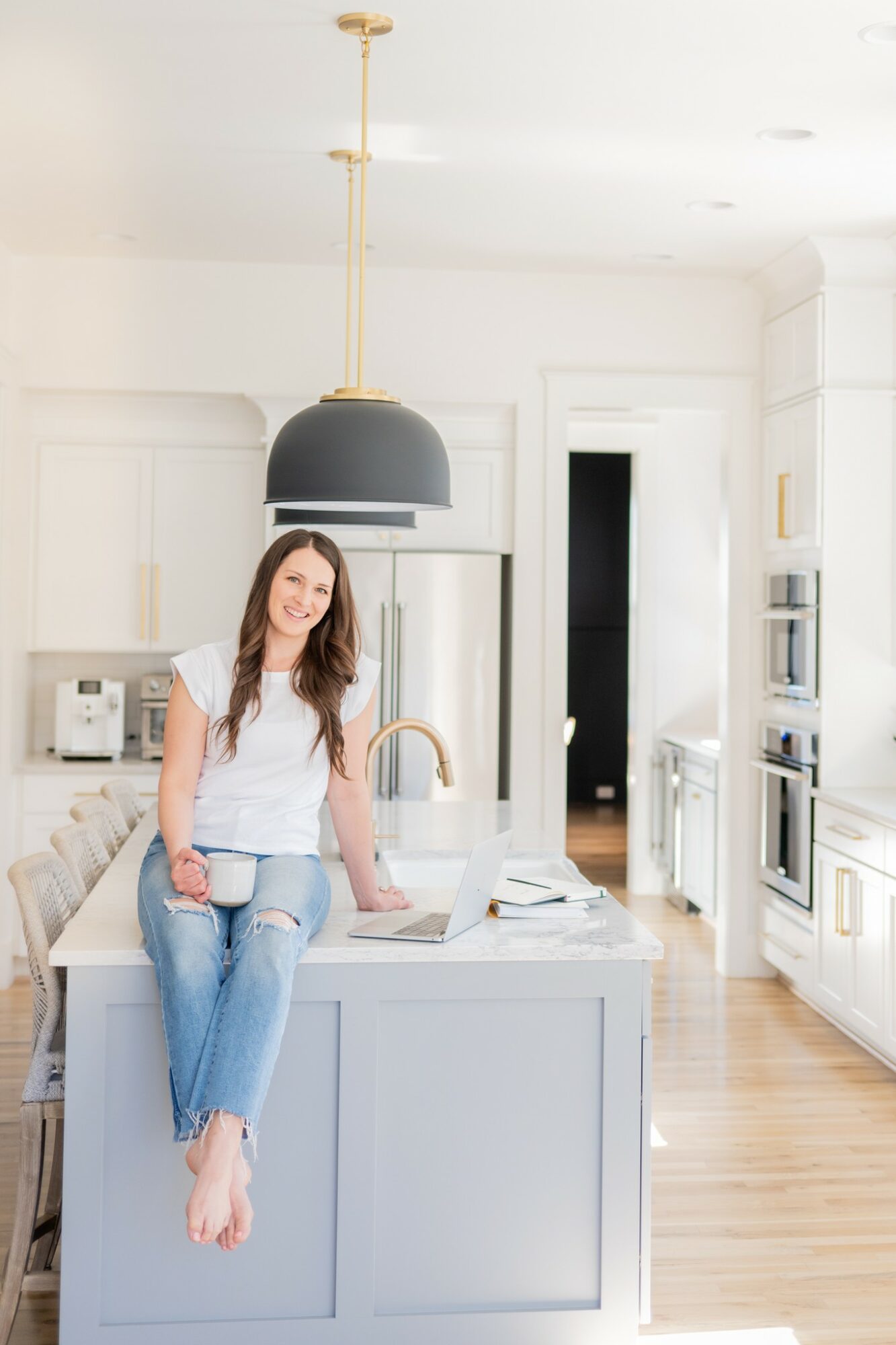 Young woman with long hair leaning on kitchen island, smiling, in a bright modern kitchen with white cabinets and black pendant light.