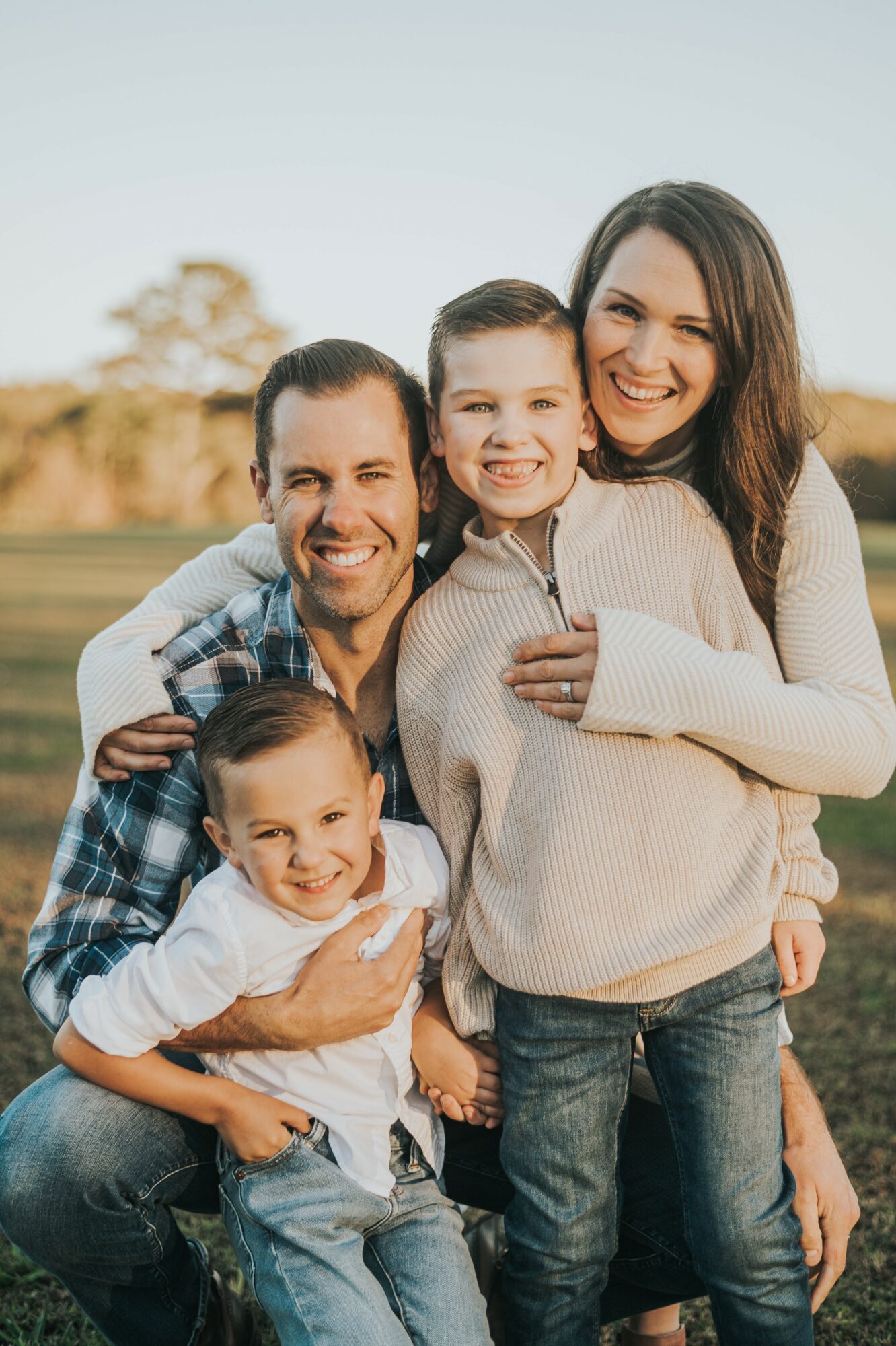 Family of four smiling outdoors, two adults and two children, standing close together, sunny day, natural background.