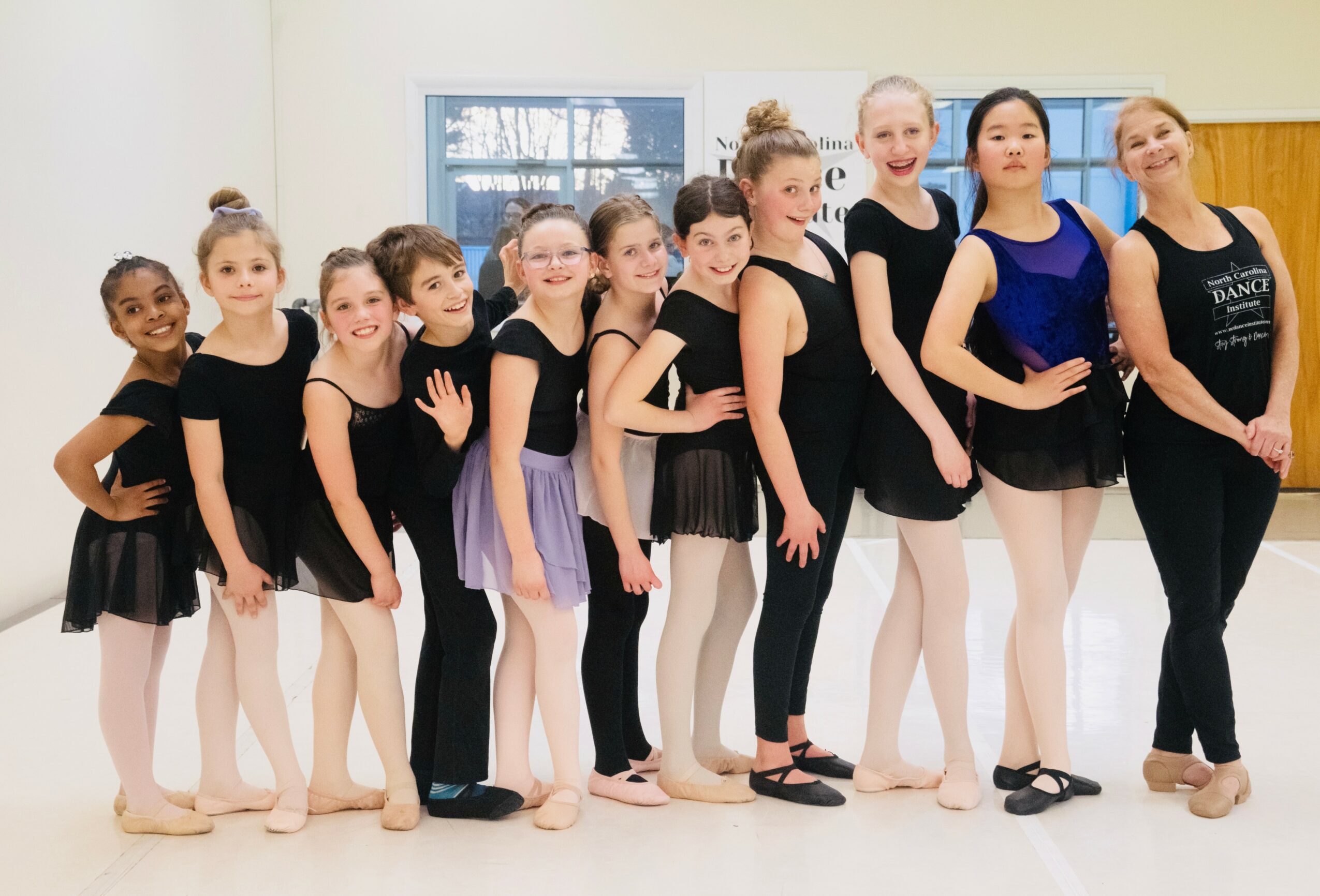 Group of young girls in dance costumes standing in a line indoors, smiling, with a window in the background.
