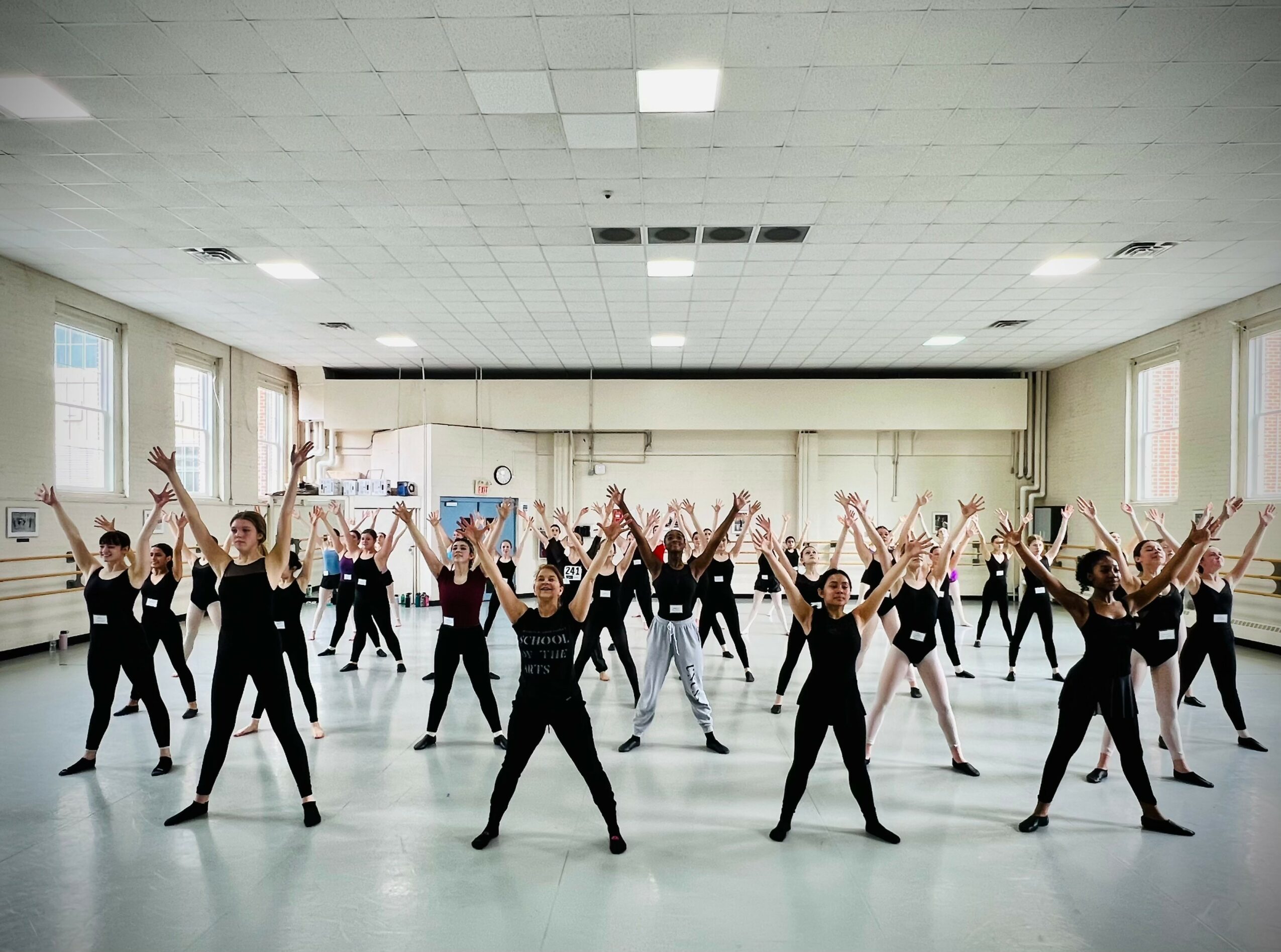 Group of people practicing dance or exercise in a spacious room with large windows, arms raised, facing forward.