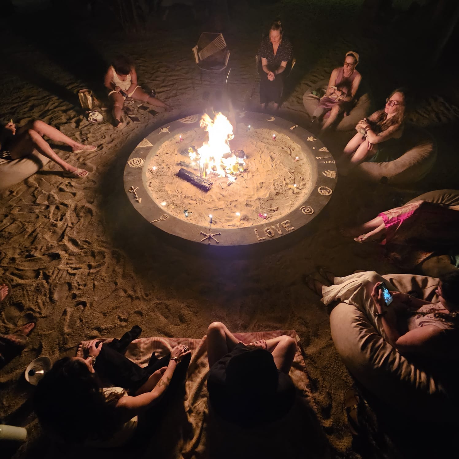 Group of people sitting around a campfire on sand at night, some sitting on chairs and others on the ground, with a woman standing nearby.