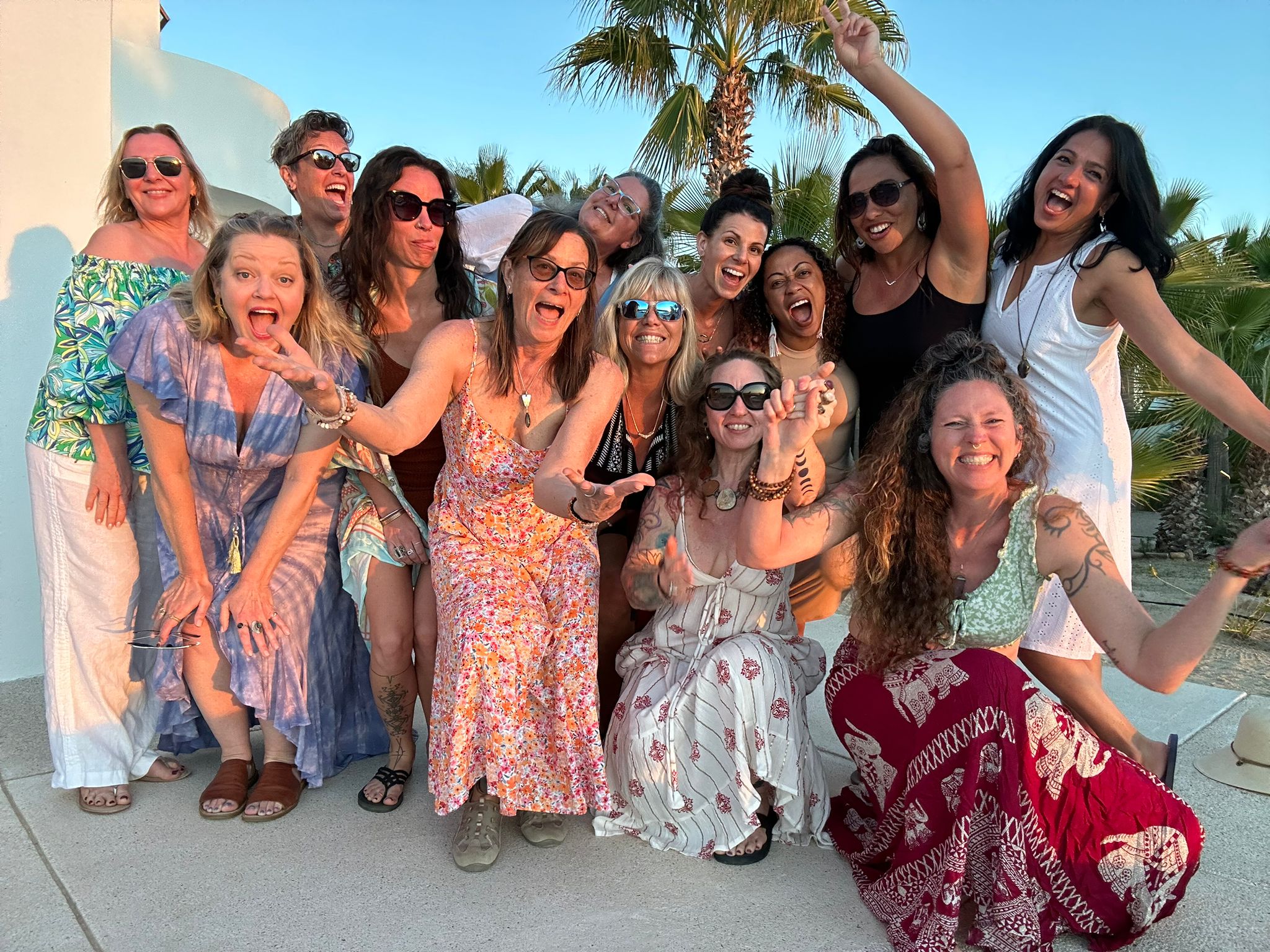 Group of women and men smiling outdoors with palm trees, wearing summer clothes, some with sunglasses, celebrating together.