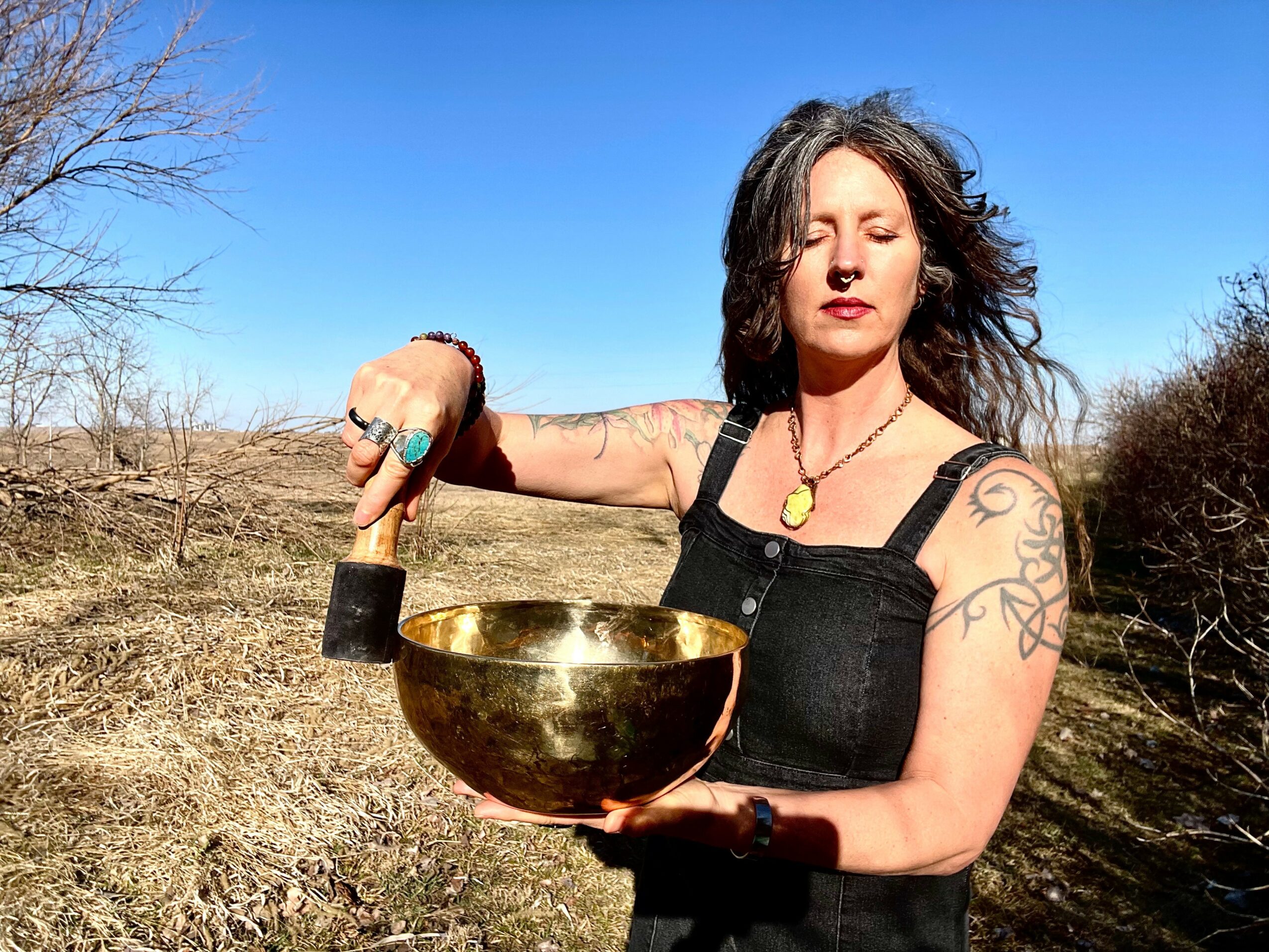 Woman outdoors holding a large singing bowl, with a clear blue sky and dry landscape in background.