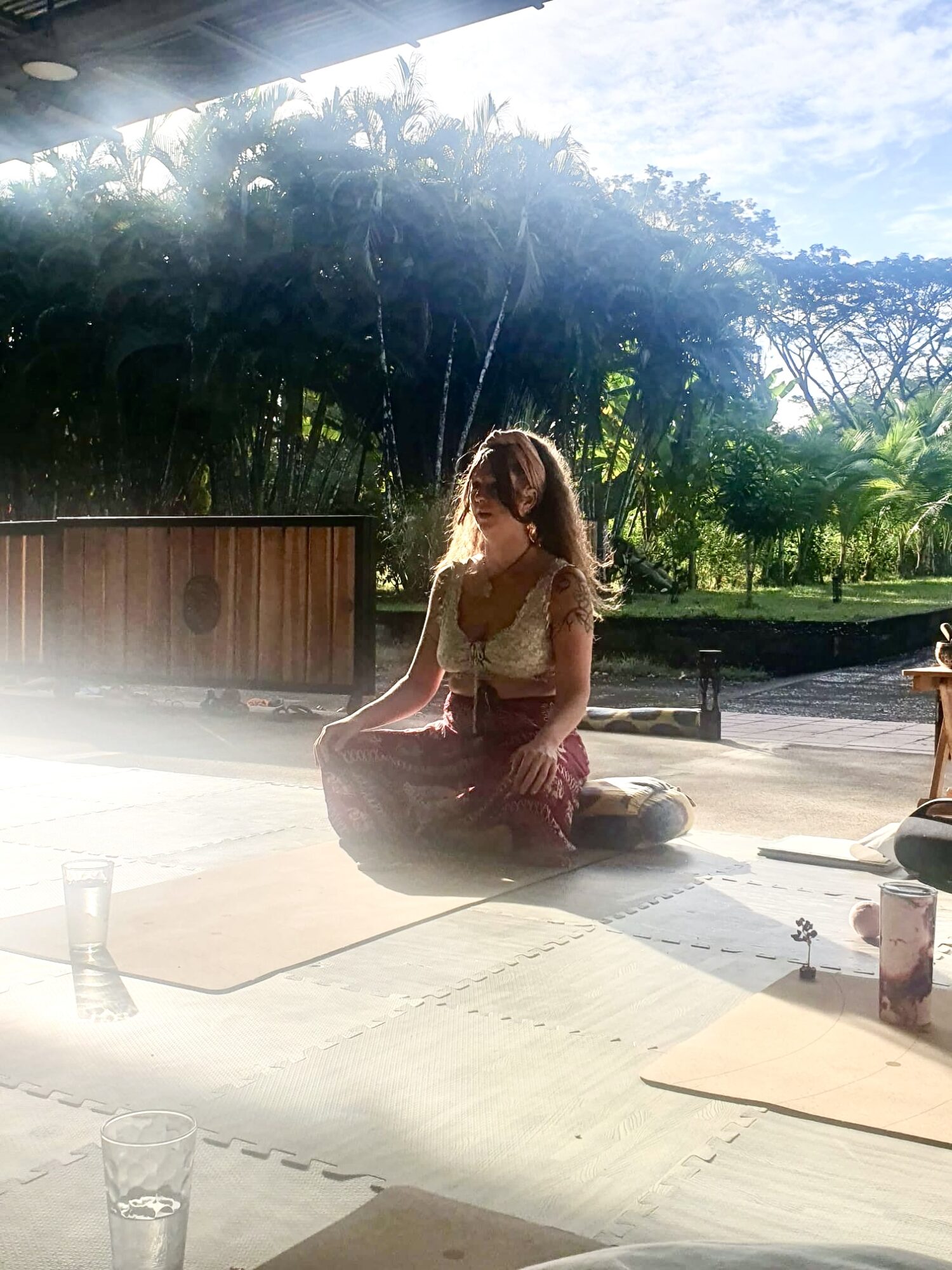 Person sitting cross-legged outdoors on a mat, with trees and sky in the background, sunlight shining on them.
