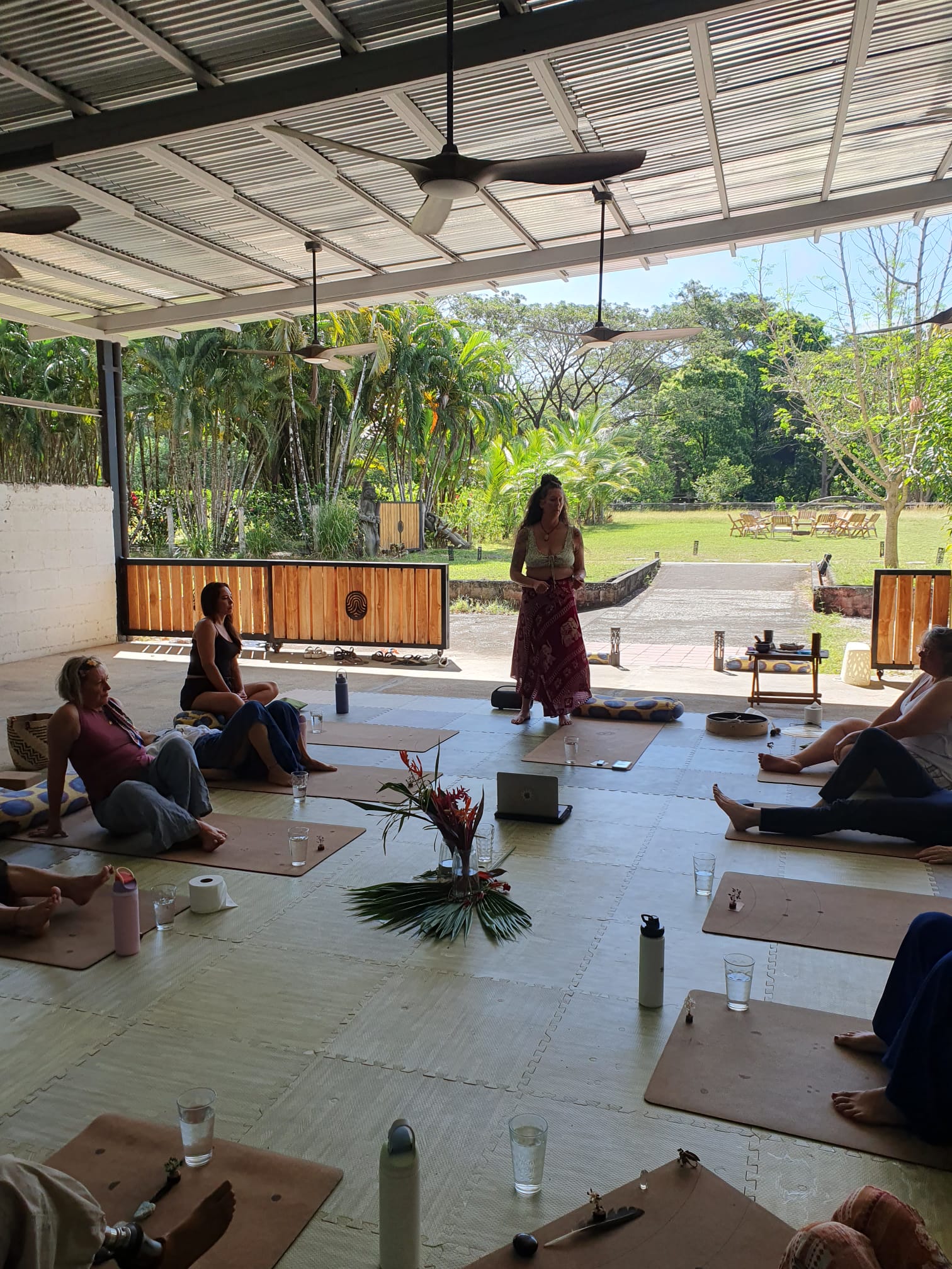 People sitting on mats and chairs in a shaded open space, with a woman standing and reading, greenery outside.