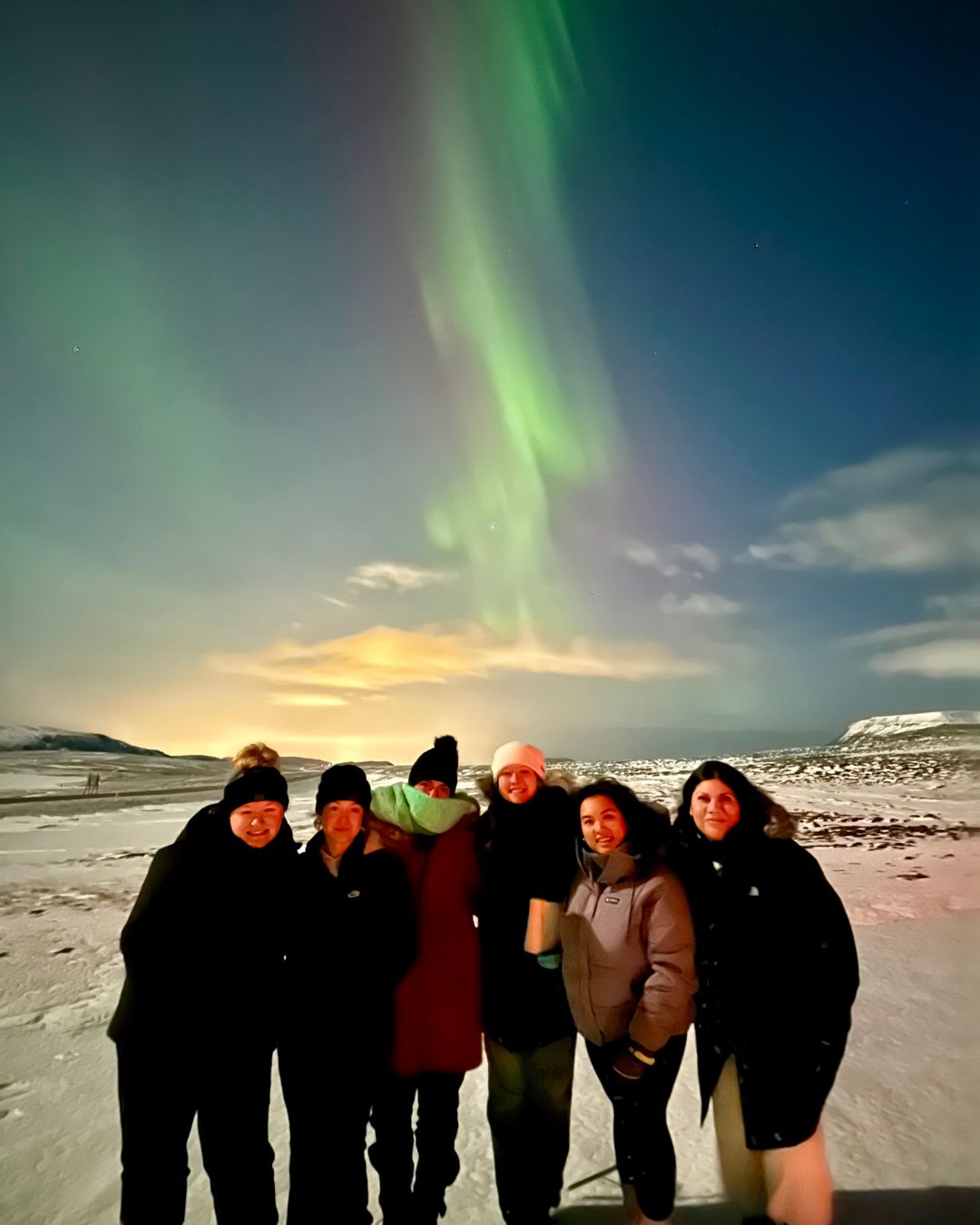 Group of five people standing outdoors at night with northern lights in the sky, snow-covered ground, and distant landscape.