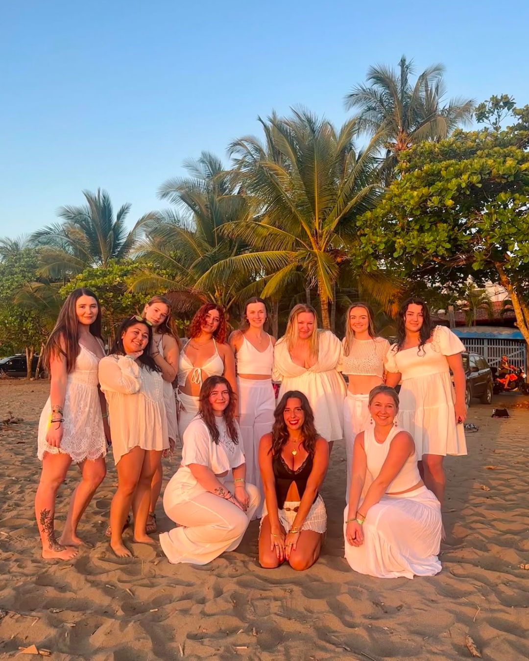 Group of ten women on a sandy beach with palm trees and blue sky, some standing and some kneeling, smiling.