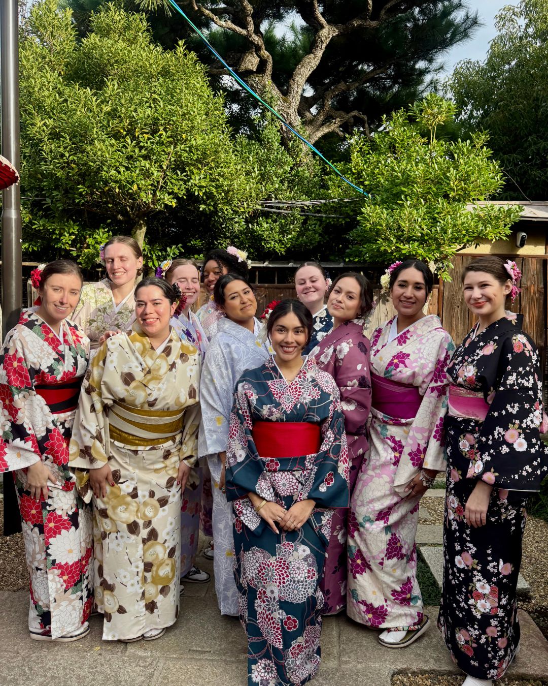 Group of women in colorful kimonos standing outdoors with trees in background.