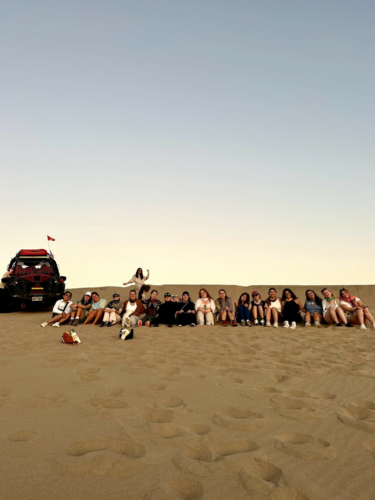 Group of people sitting on sand dune with a vehicle and flag in the background, under a clear sky.