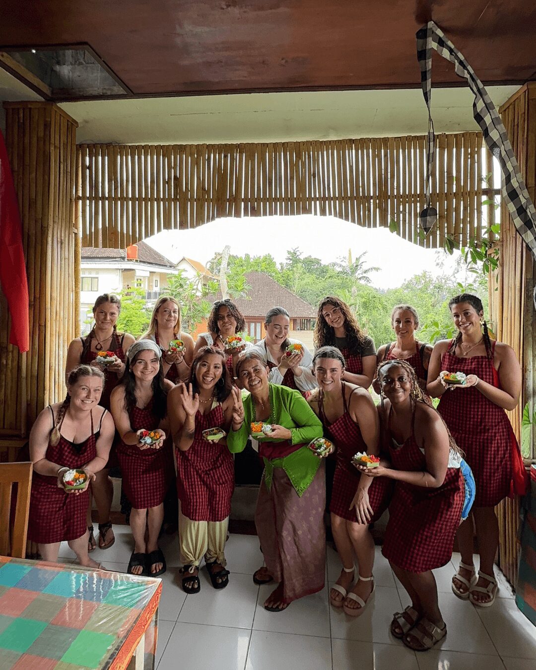 Group of women in traditional dresses posing indoors with a bamboo wall and outdoor view behind them.
