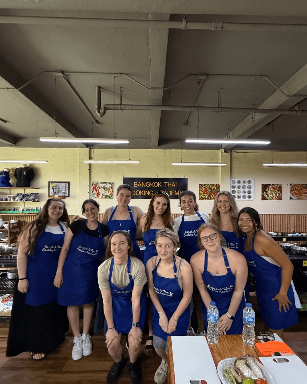 Group of ten women wearing blue aprons inside a store, some smiling, with shelves and artwork in the background.