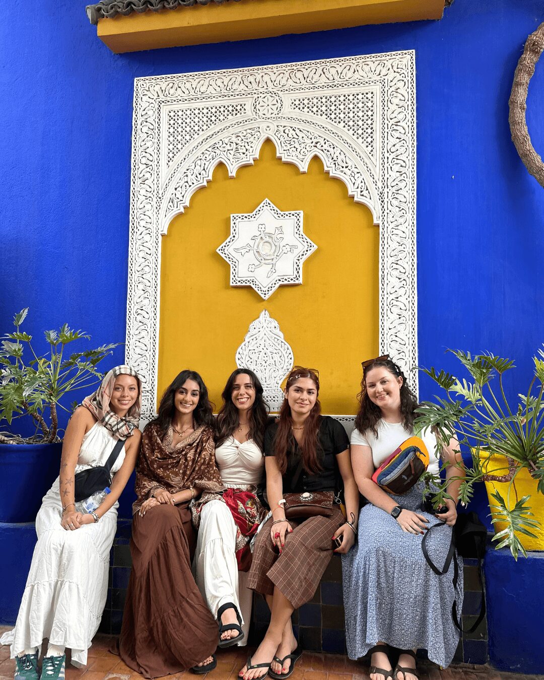 Four women sitting on a bench in front of a colorful wall with intricate white arch design, plants on sides, and a yellow section.