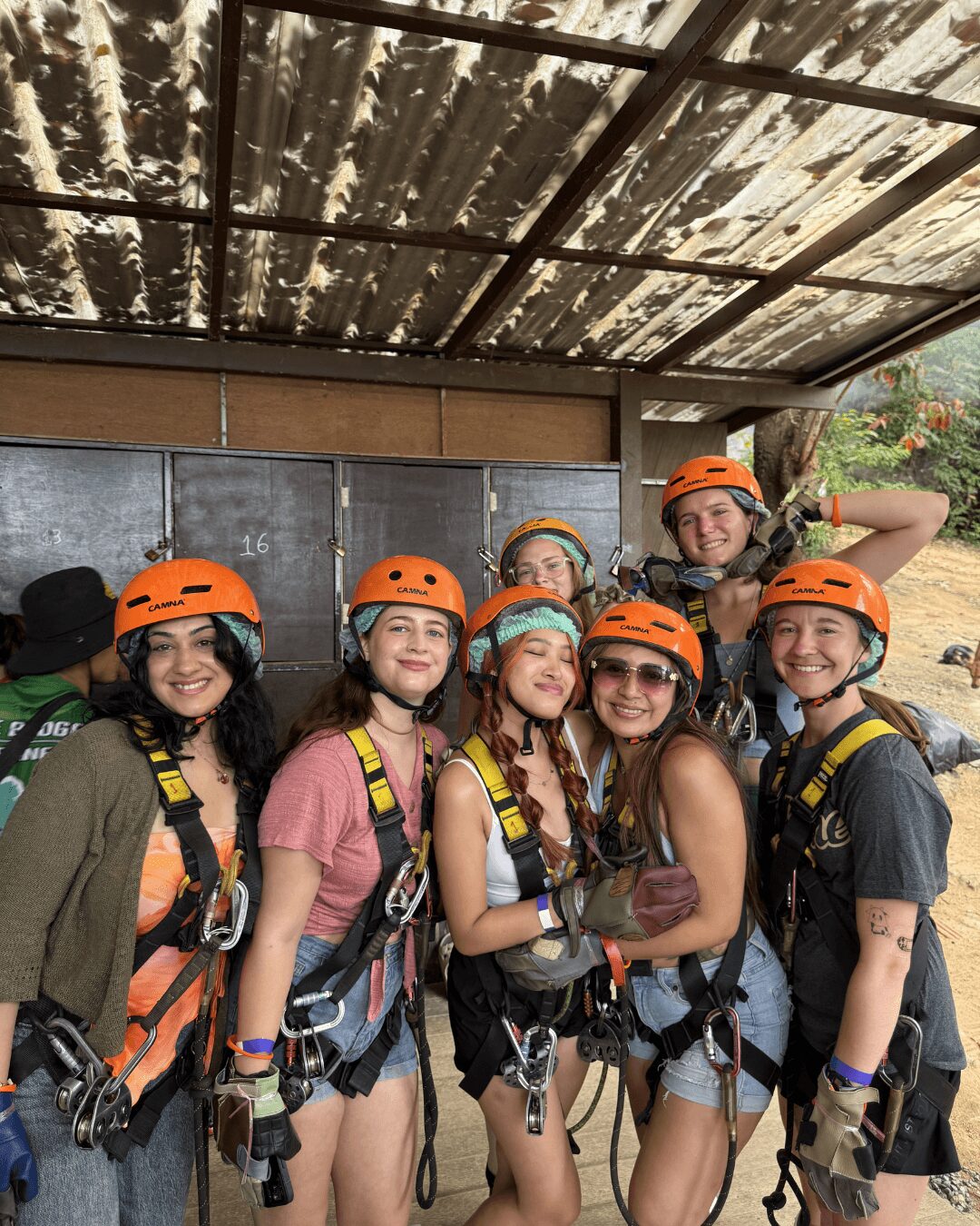 Group of six young women wearing helmets and harnesses, standing outdoors under a metal roof, smiling at the camera.