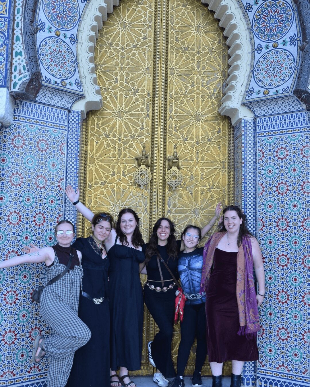 Five women stand in front of an ornate, gold-colored door with intricate blue and white tile patterns surrounding it.
