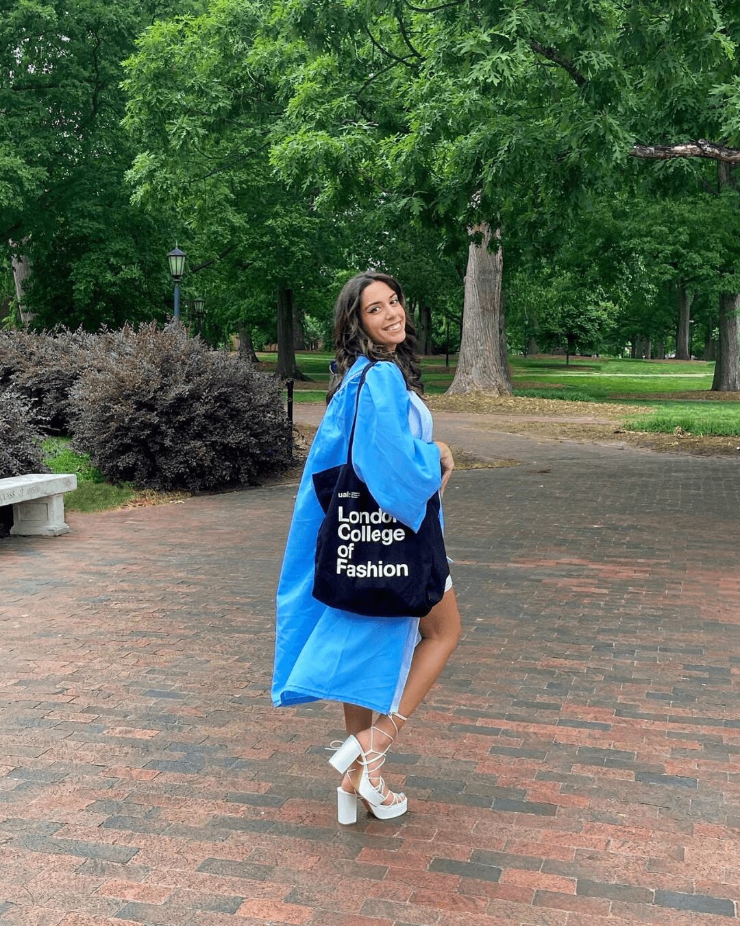 Young woman in a blue coat and white shoes standing on a brick path with trees and greenery in the background.