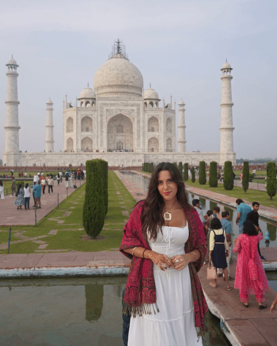 Woman in white dress and pink shawl standing in front of the Taj Mahal with visitors nearby.